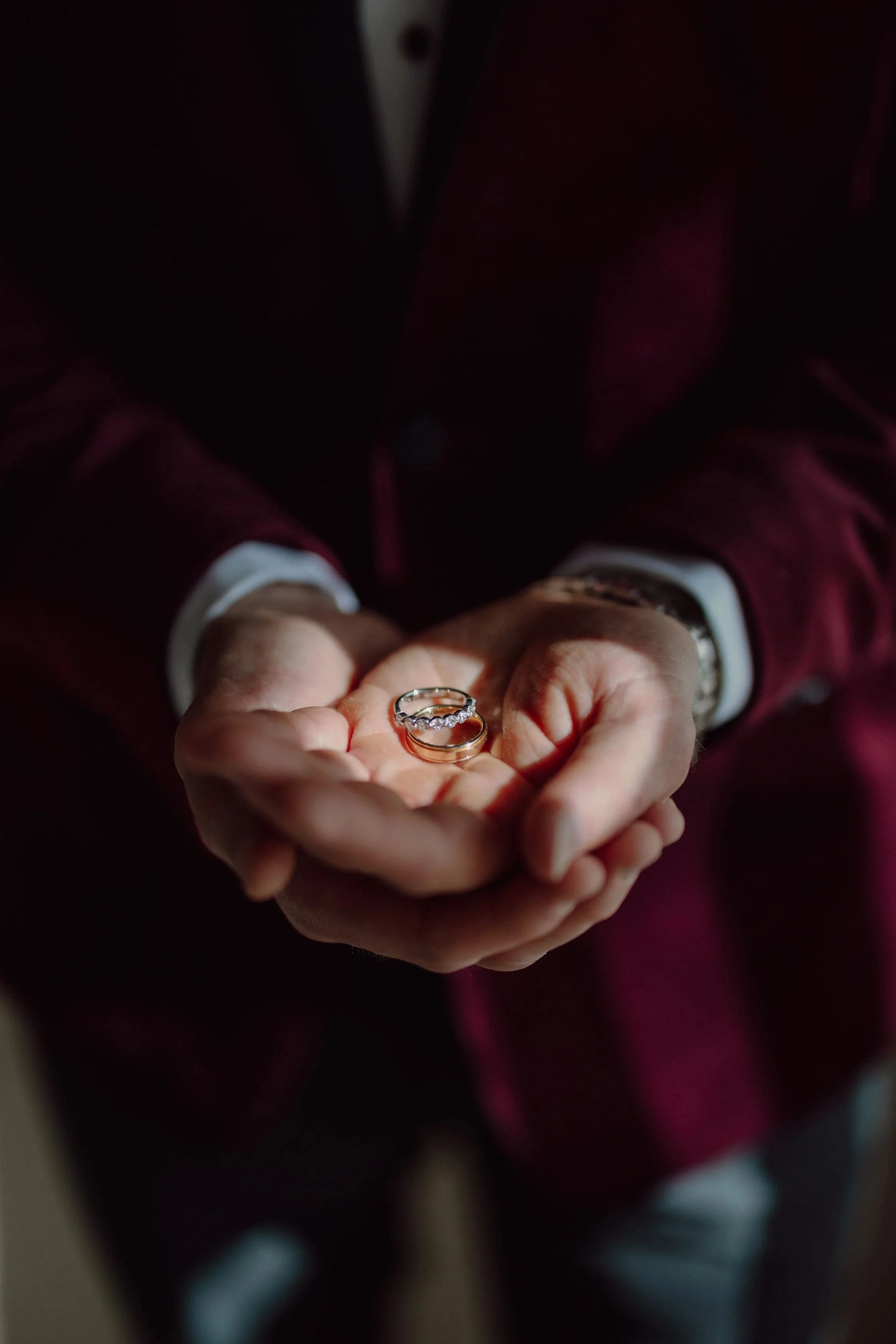 Anthony holds both wedding rings in his cupped hands, a beam of light catching the gold against his burgundy velvet jacket.