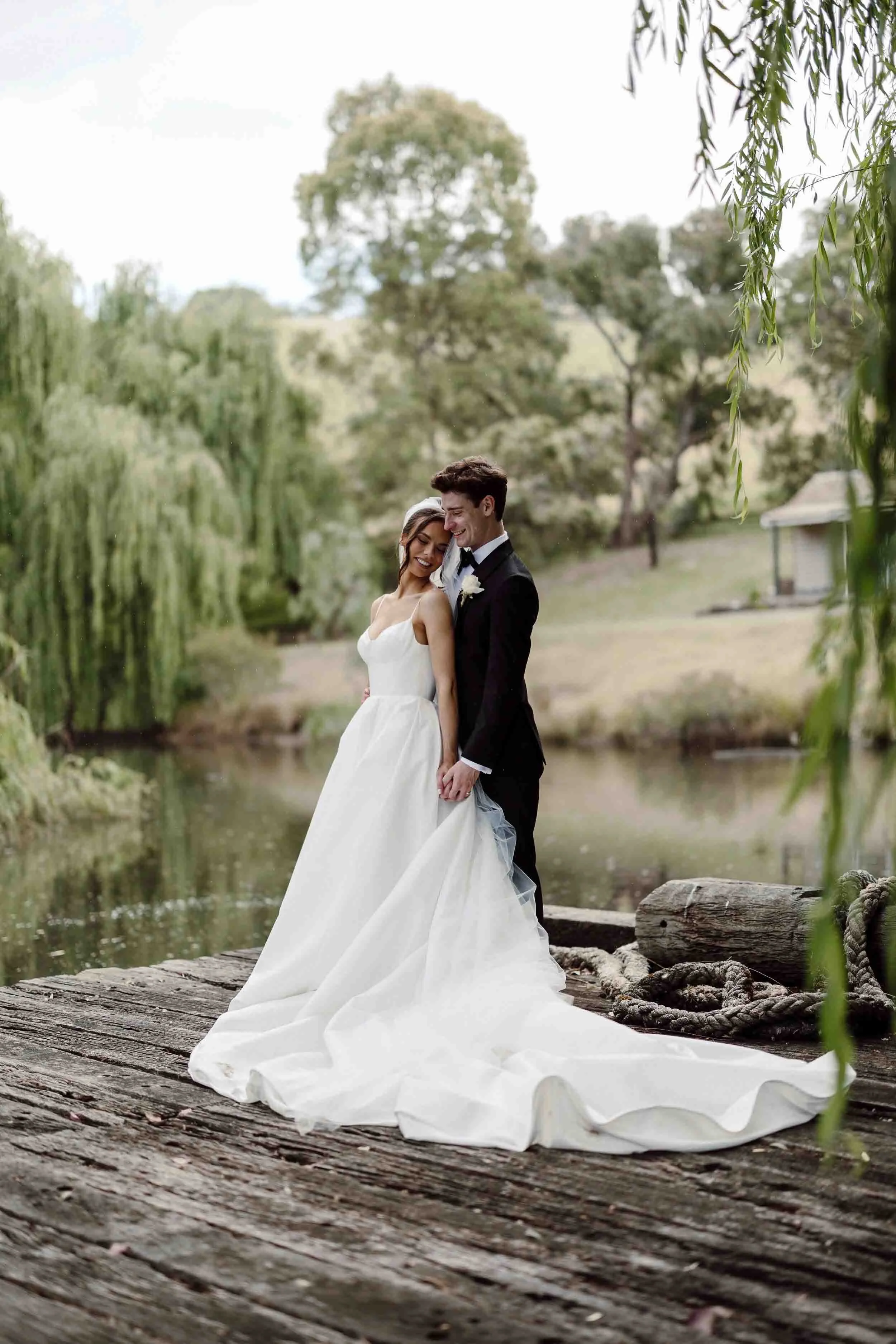 Ruby rests against Jacob on the timber jetty at Inglewood Estate, willows and still water behind them.
