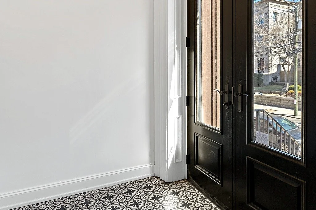 Historic townhouse doorway with glass panels, patterned tile floor, and sunlight entering from outside.