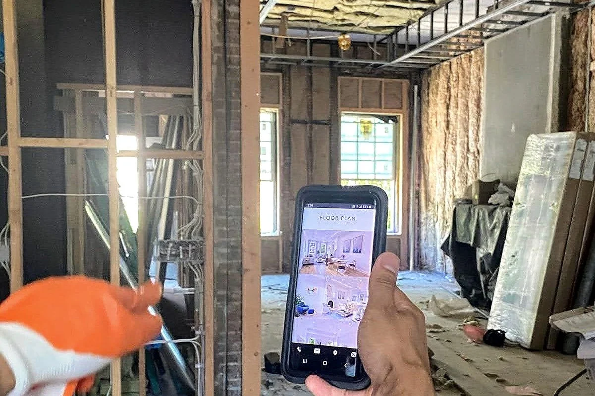 Construction site interior with exposed framing and insulation as a floor plan is viewed on a phone.