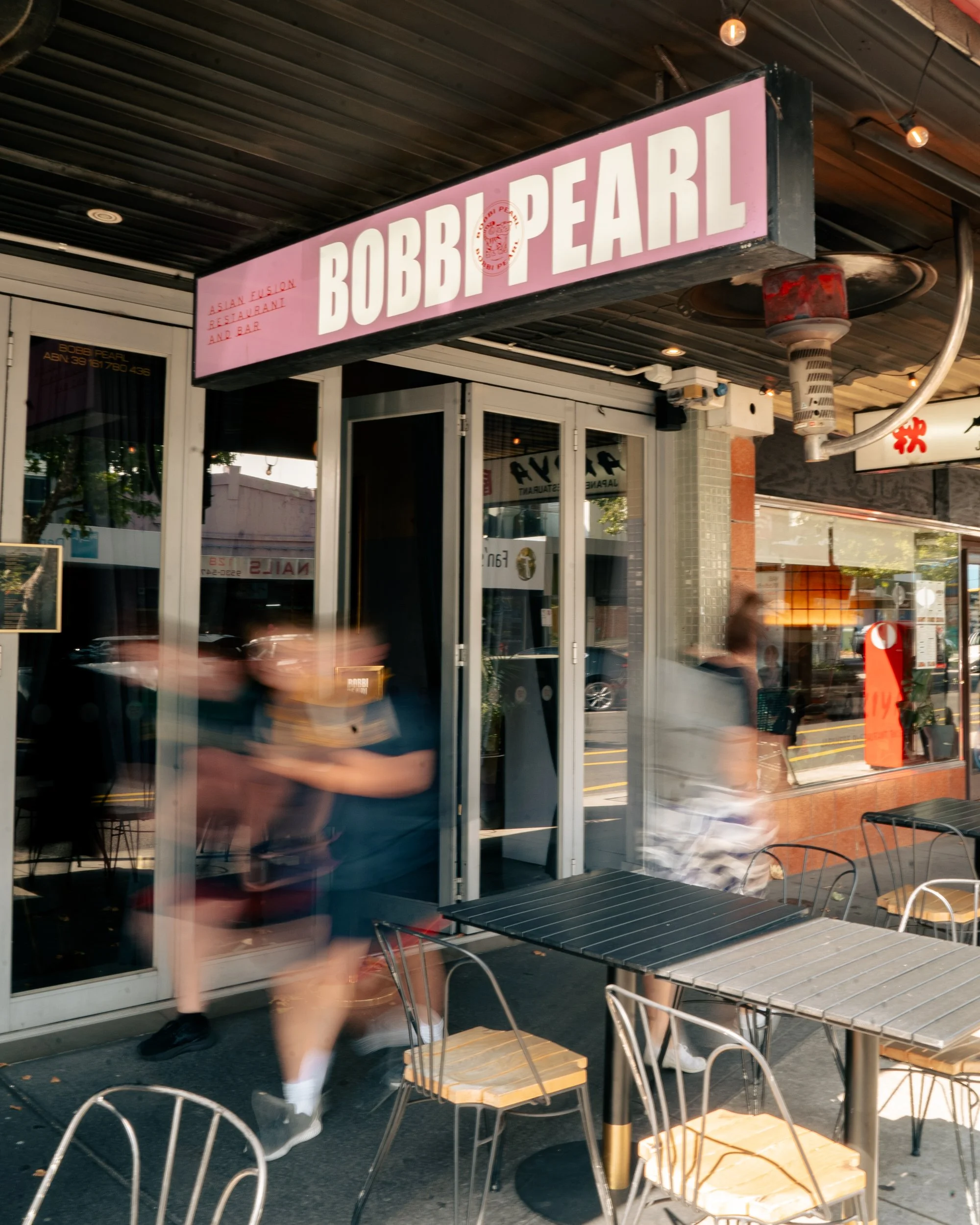 The exterior of Bobbi Pearl, an Asian fusion restaurant and bar, featuring a pink illuminated sign, outdoor seating with metal tables and chairs, and blurred pedestrians walking past on the sidewalk.