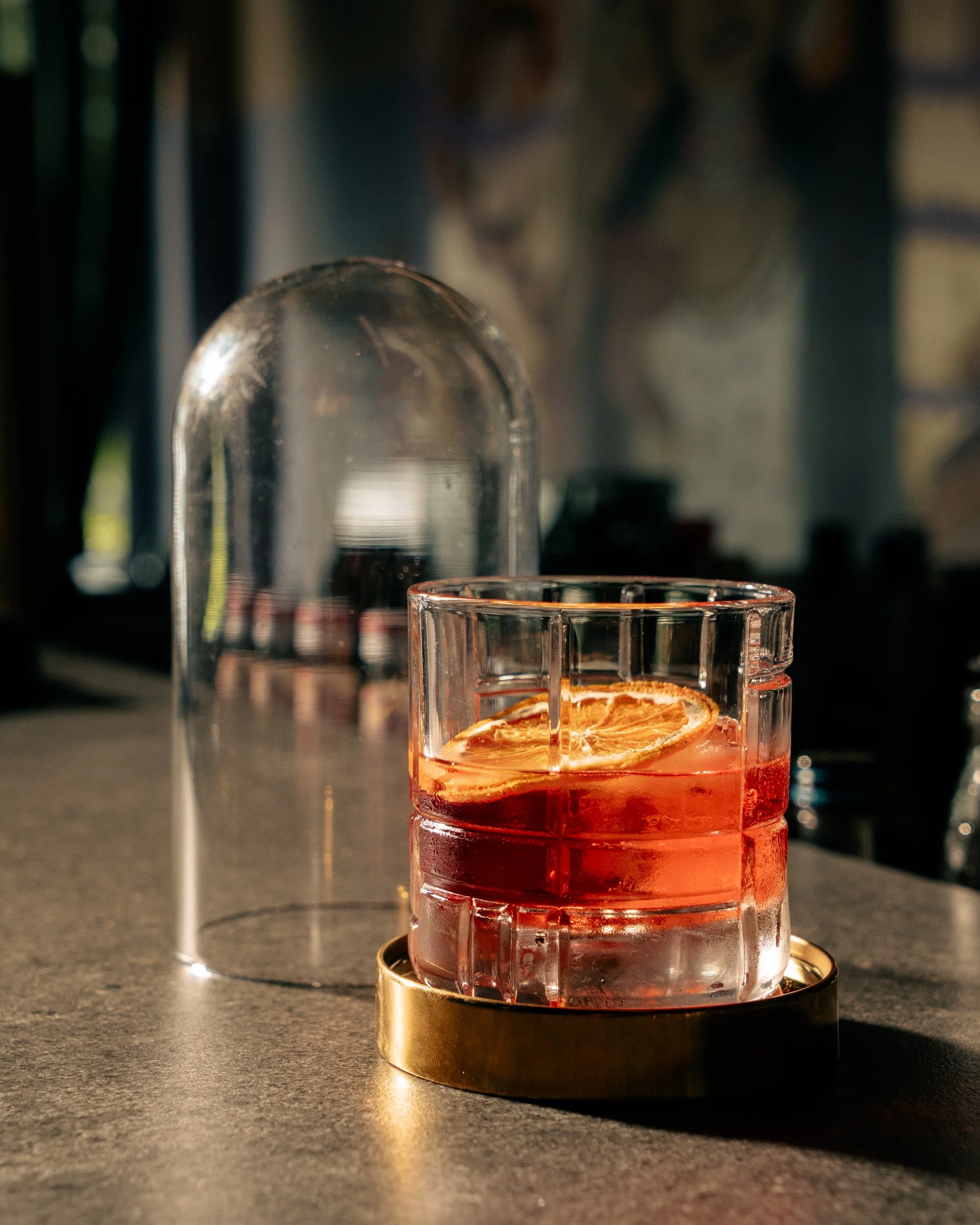 A glass of red cocktail with lemon slice garnis, sitting on a gold coaster on a bar counter, with a glass dome in the background.