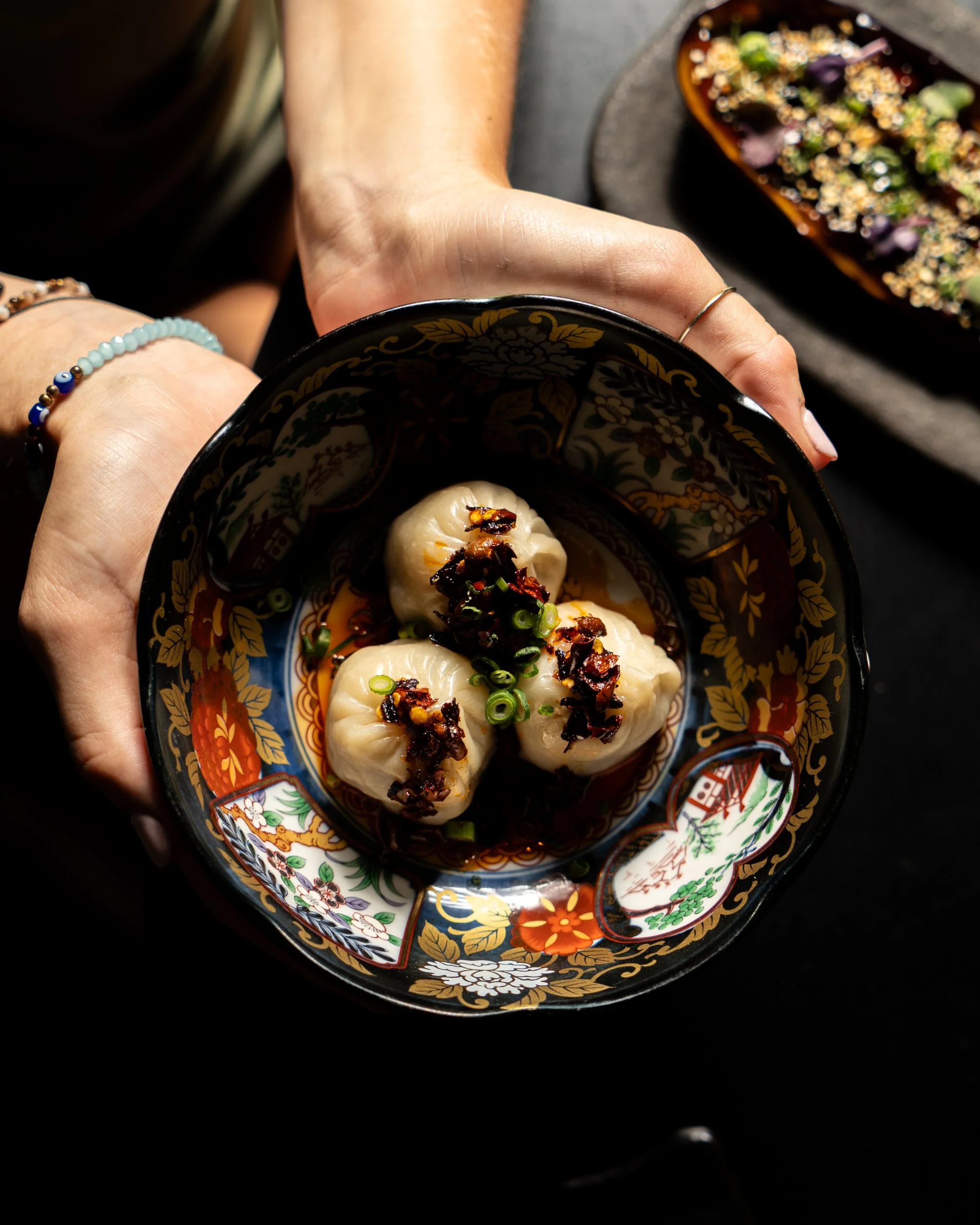 A hand holding a decorative black bowl with three Asian-style dumplings topped with chopped green onions and crispy fried garlic, with a second dish of condiments or garnishes on a dark surface in the background.