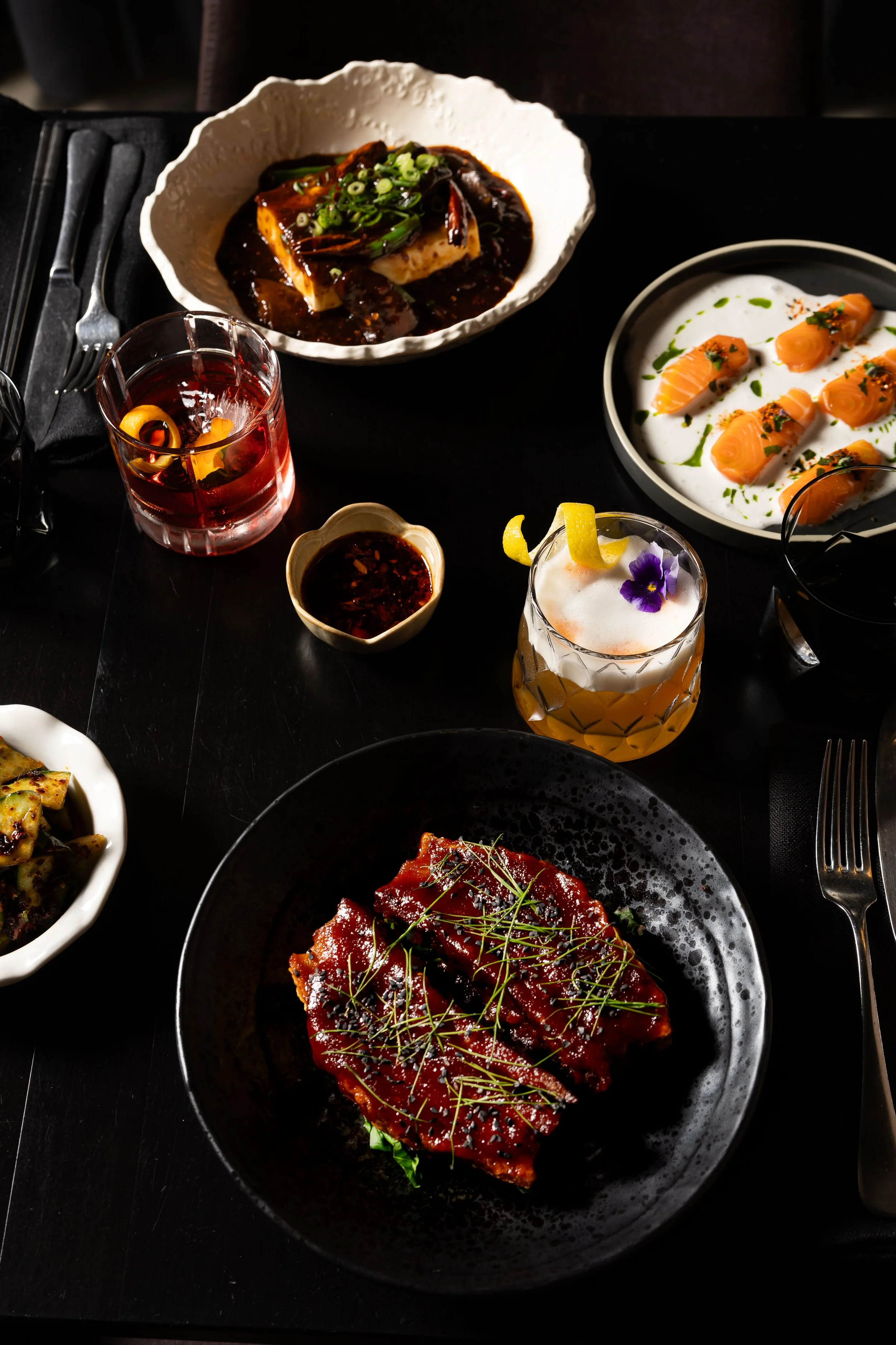 Assorted Asian dishes including sashimi, glazed meat, tofu with sauce, and several beverages on a black table.
