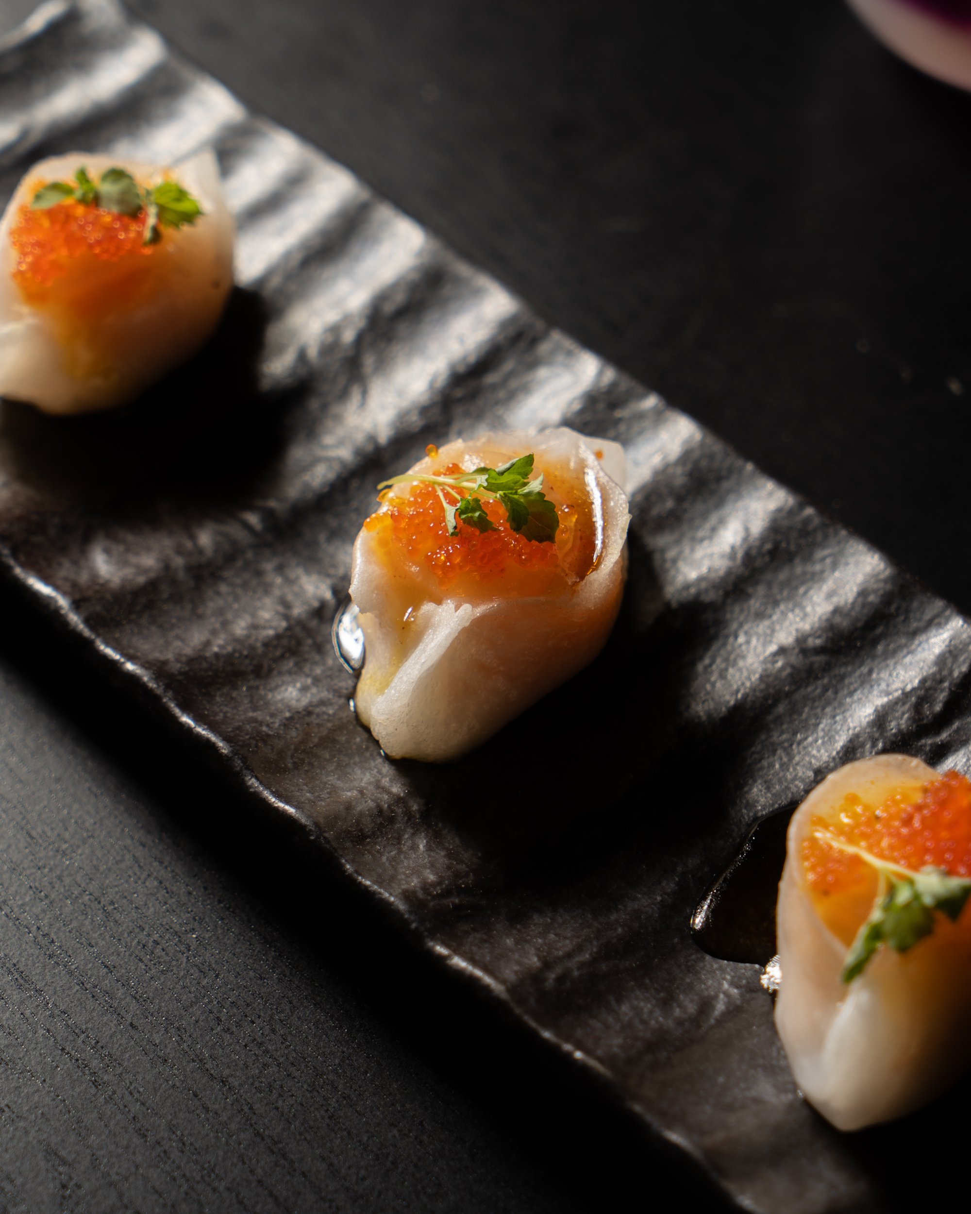 Close-up of three pieces of sushi with white rice, fish, orange fish roe, and green herbs on a textured black plate.