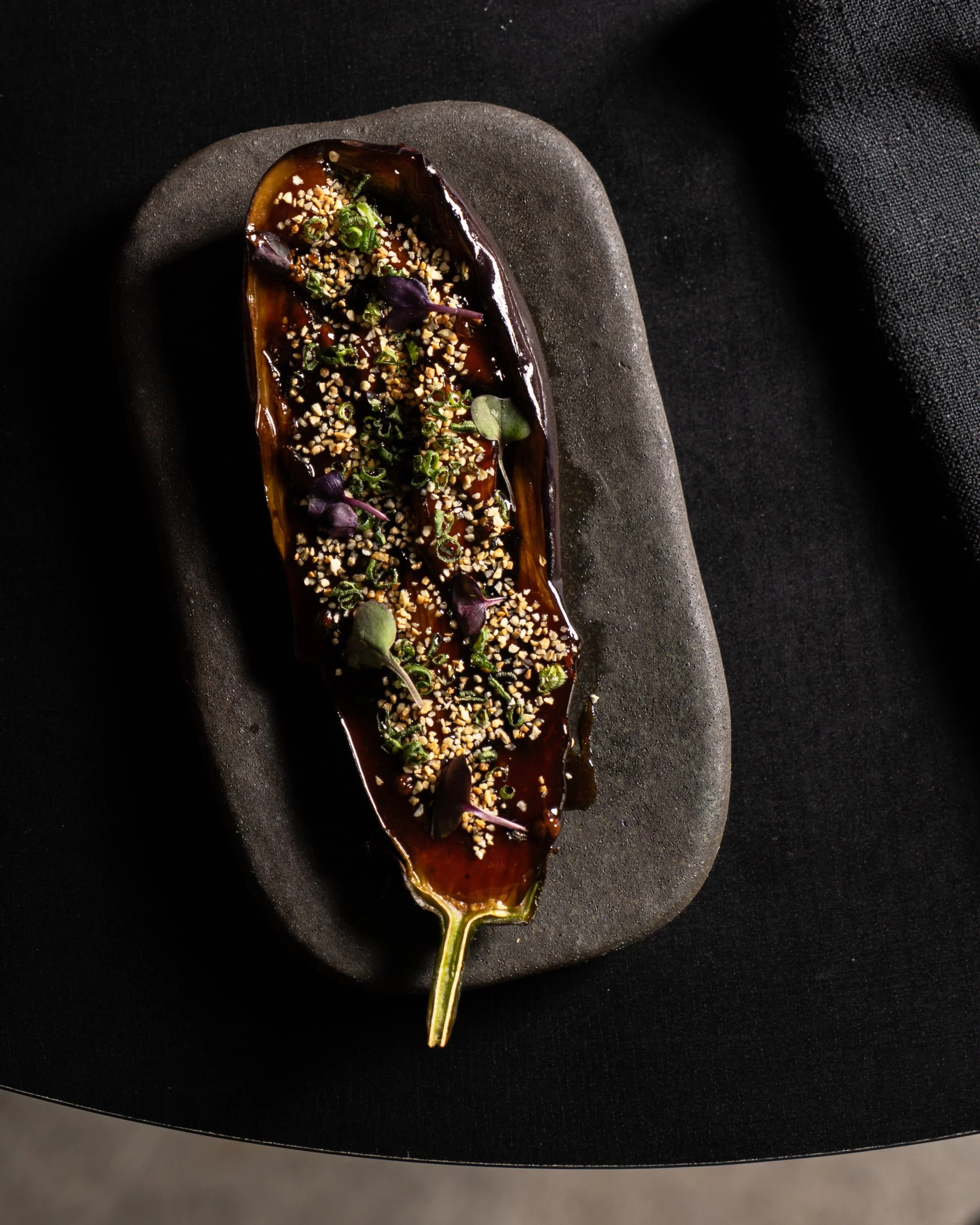 Roasted eggplant topped with chopped nuts, microgreens, and purple edible flowers on a black plate.