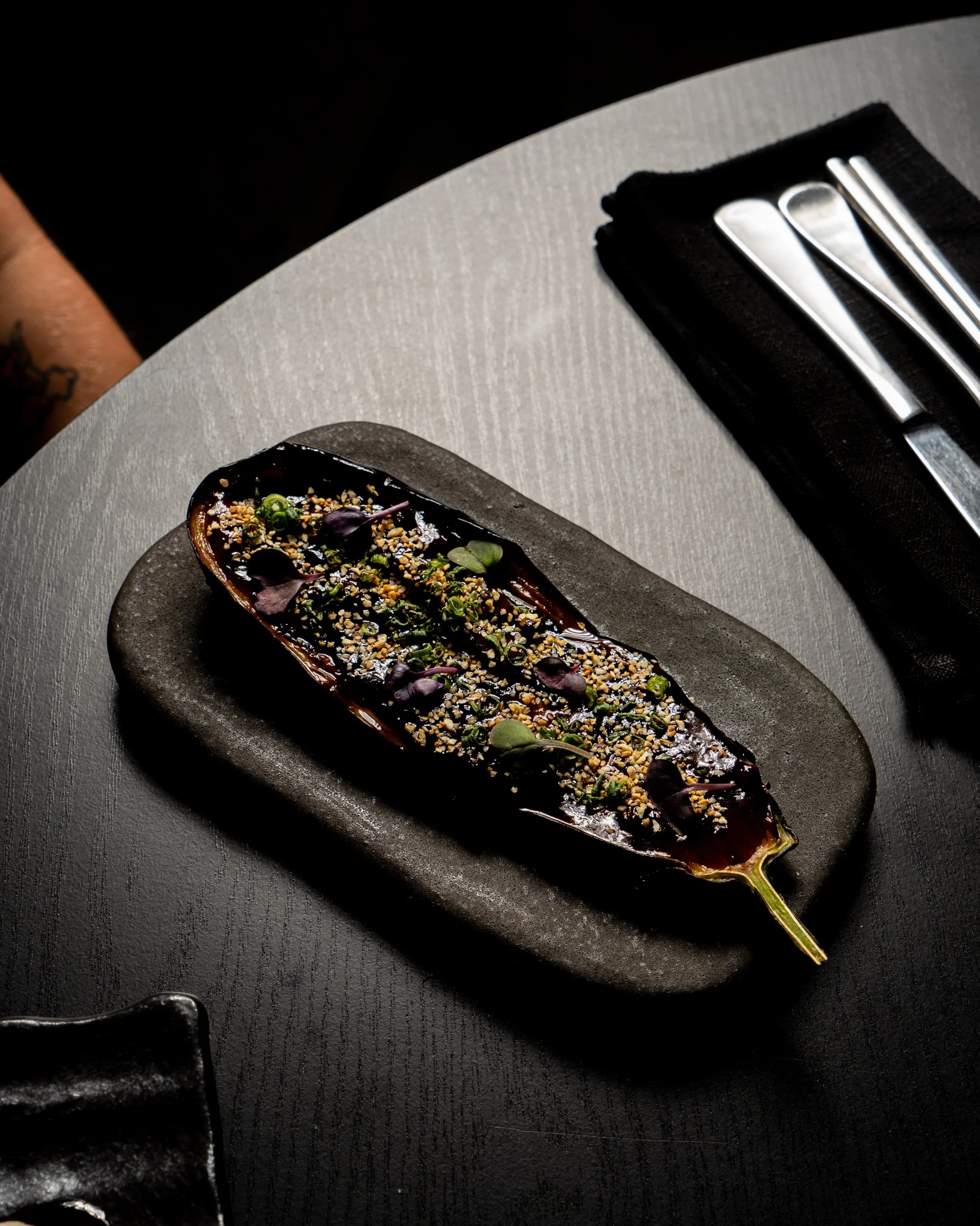 A gourmet dish with a long roasted eggplant topped with microgreens, sesame seeds, and other garnishes, served on a dark, oval plate on a textured gray table.