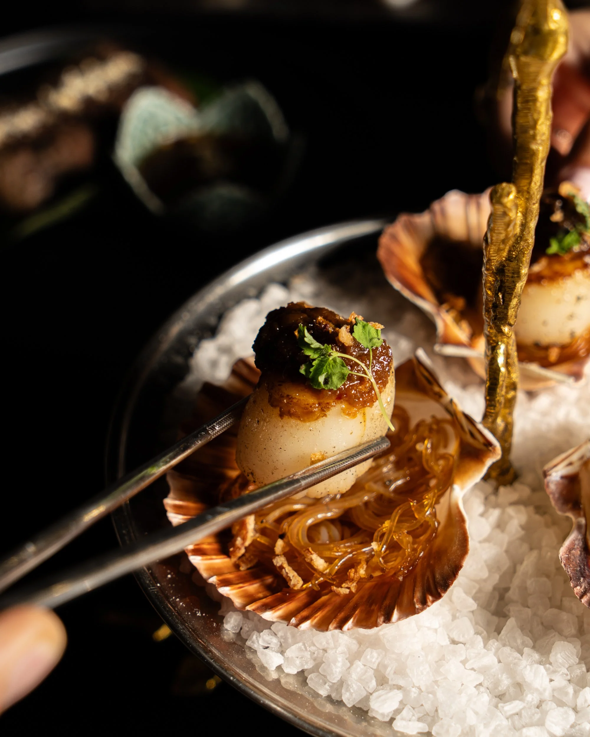 Close-up of a gourmet seafood dish featuring shellfish on ice with garnishes, being delicately handled with metal tongs.