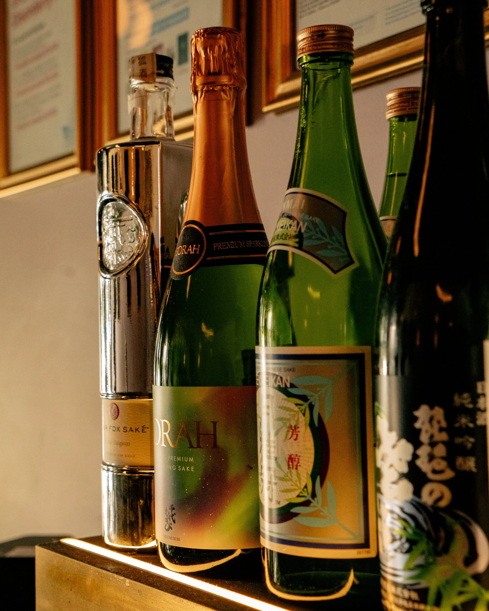 Various bottles of sake on display, including green and black bottles, some with gold and black labels, arranged on a lit shelf.