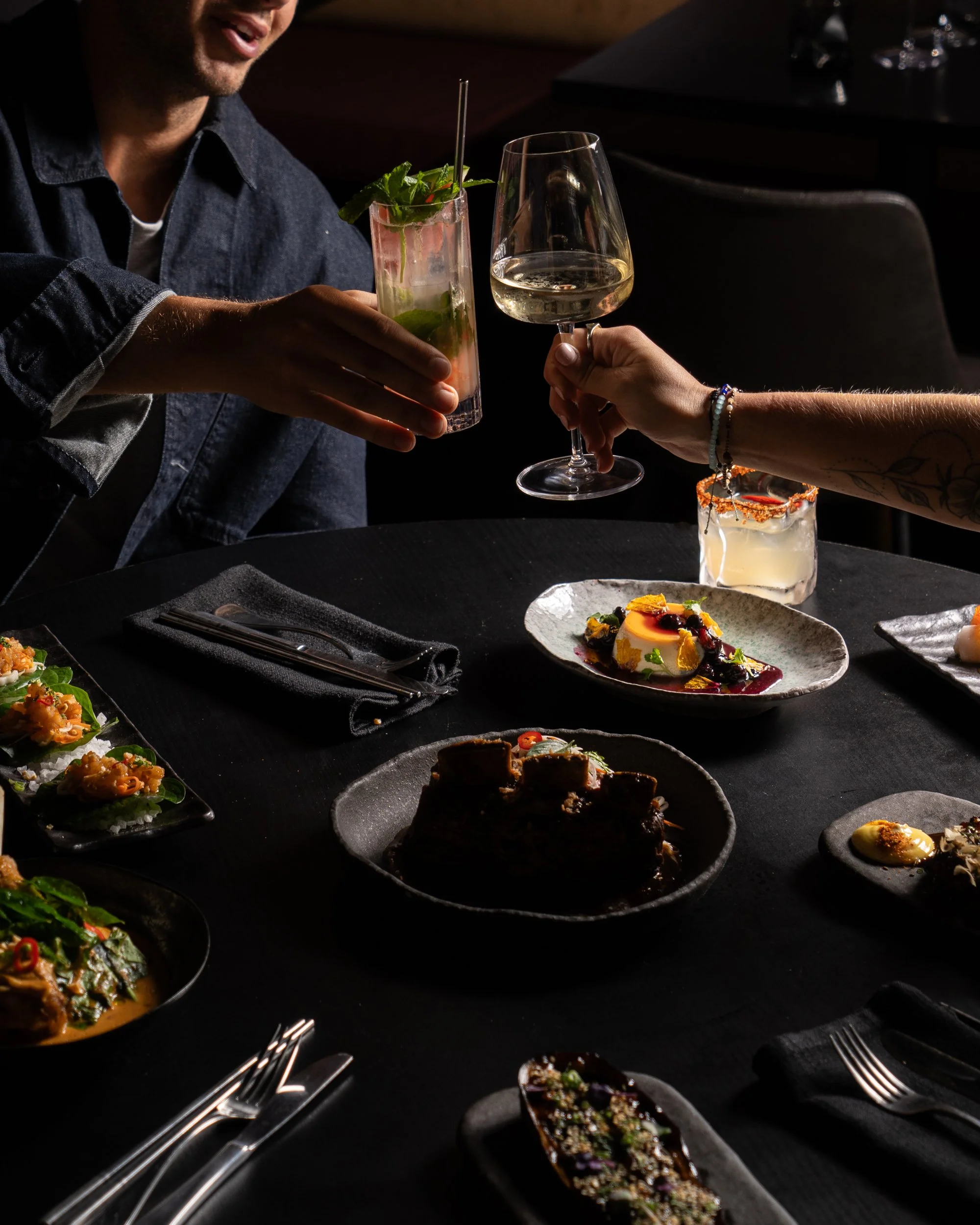People sharing drinks at a dinner table with various plated dishes, including desserts, in a dimly lit setting.