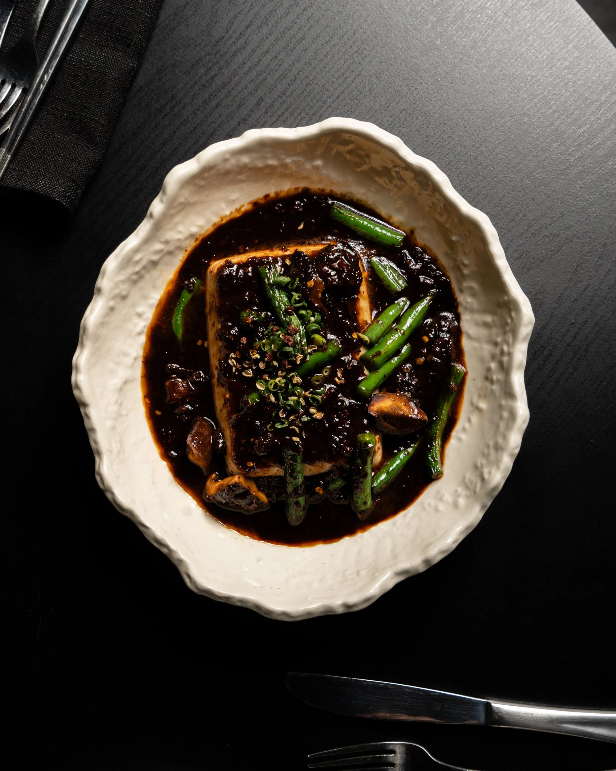 A bowl of tofu topped with green onions, sesame seeds, and a dark, savory sauce with vegetables, on a black table with utensils nearby.
