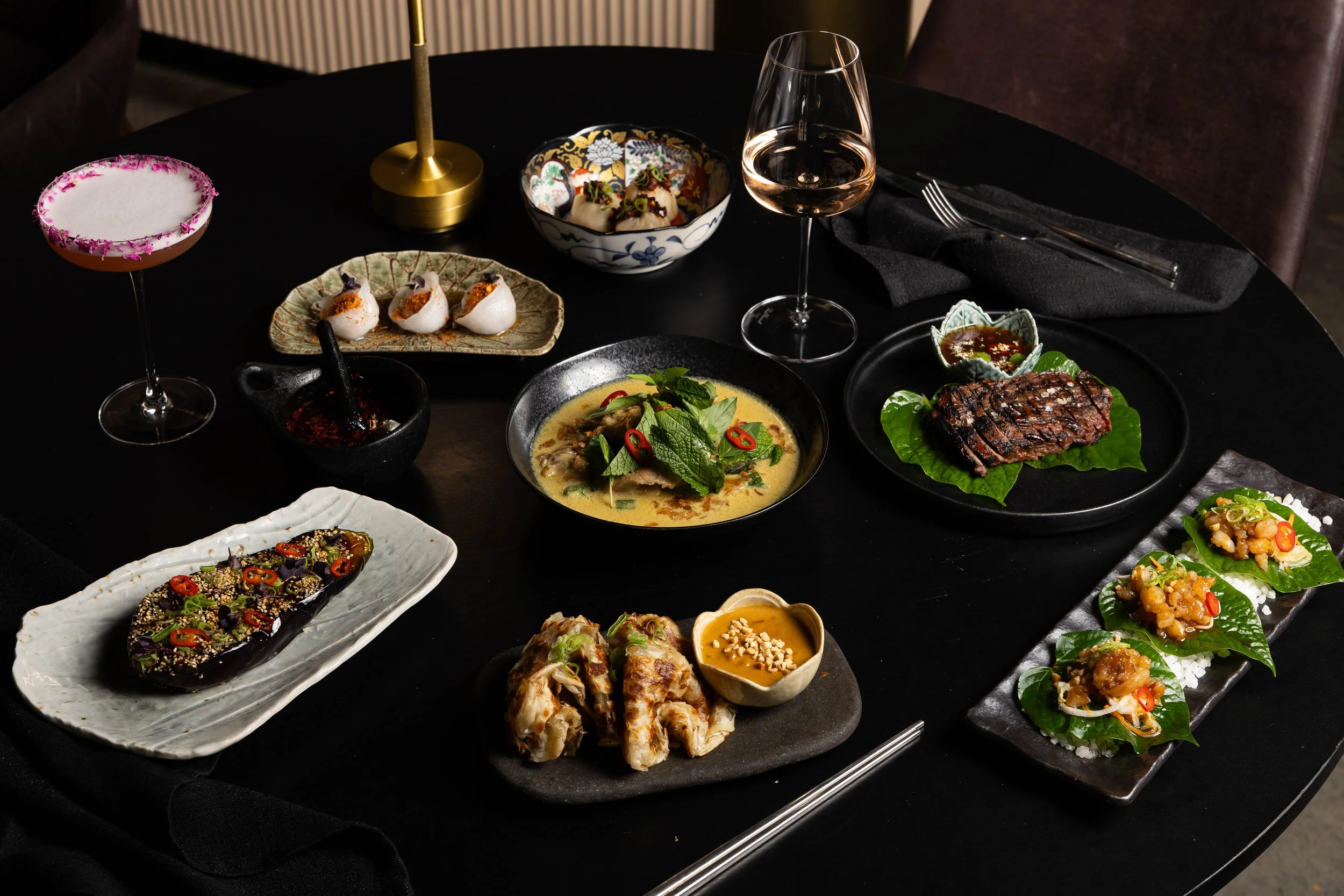 A black round table set with various Asian dishes, including a bowl of soup, grilled meat, sushi, and appetizers, with a glass of rosé wine, chopsticks, and decorative candle.