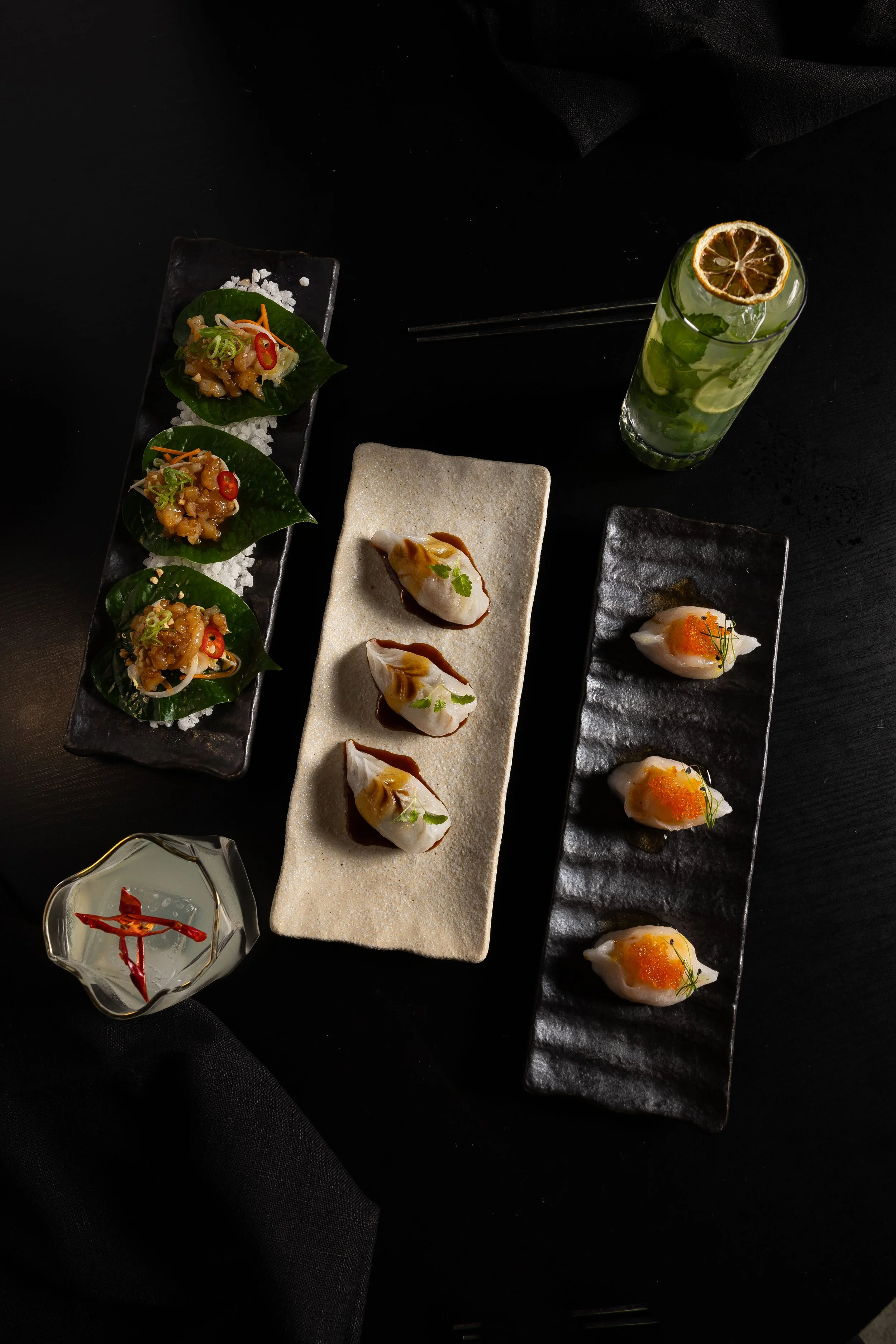 Assorted Japanese sushi and appetizers arranged on black rectangular plates with a glass of green cocktail garnished with a slice of lemon and cucumber, set on a black table.