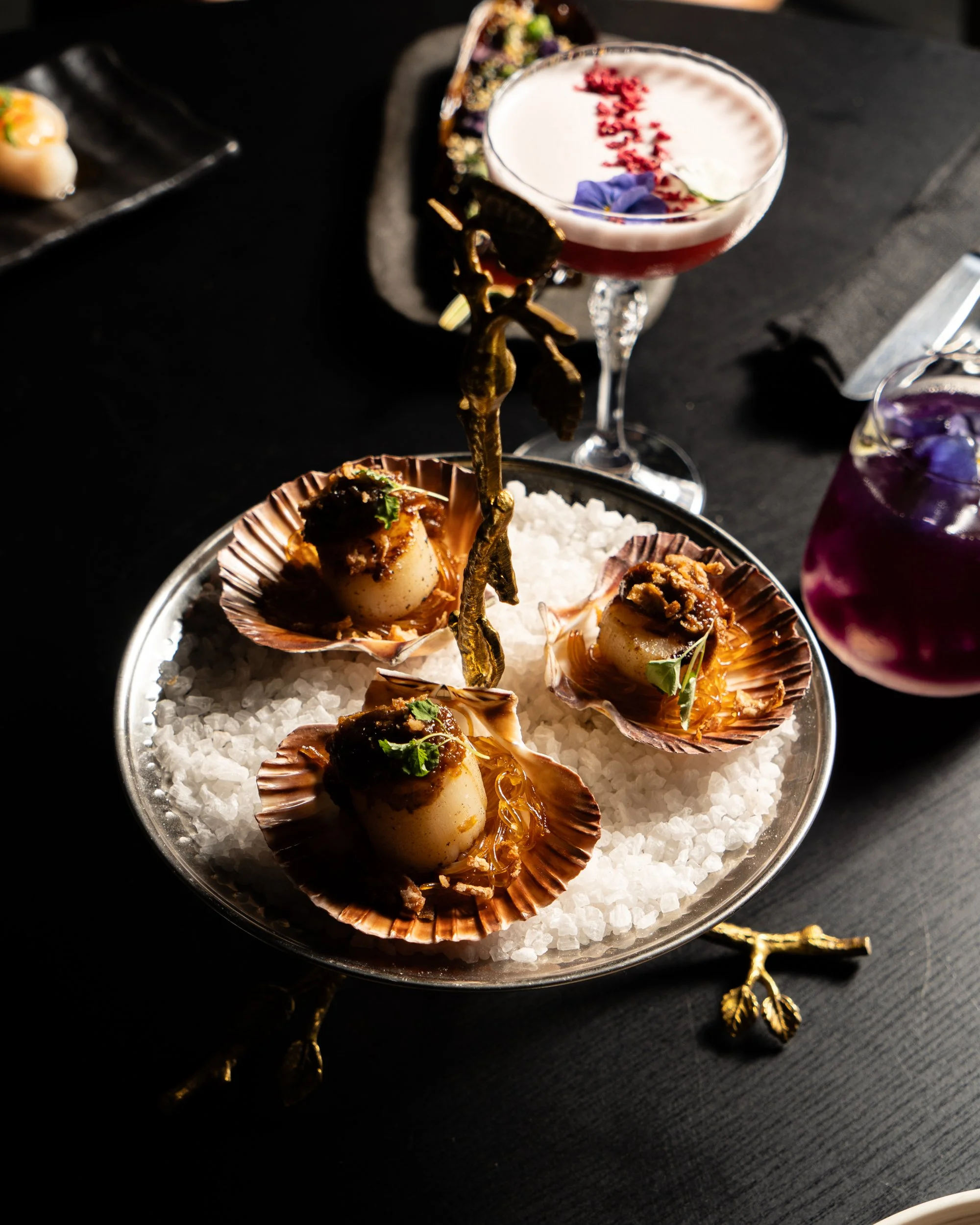 Seafood dish with scallops served in shells on a bed of ice, garnished with herbs and sauce, with colorful cocktails in the background.