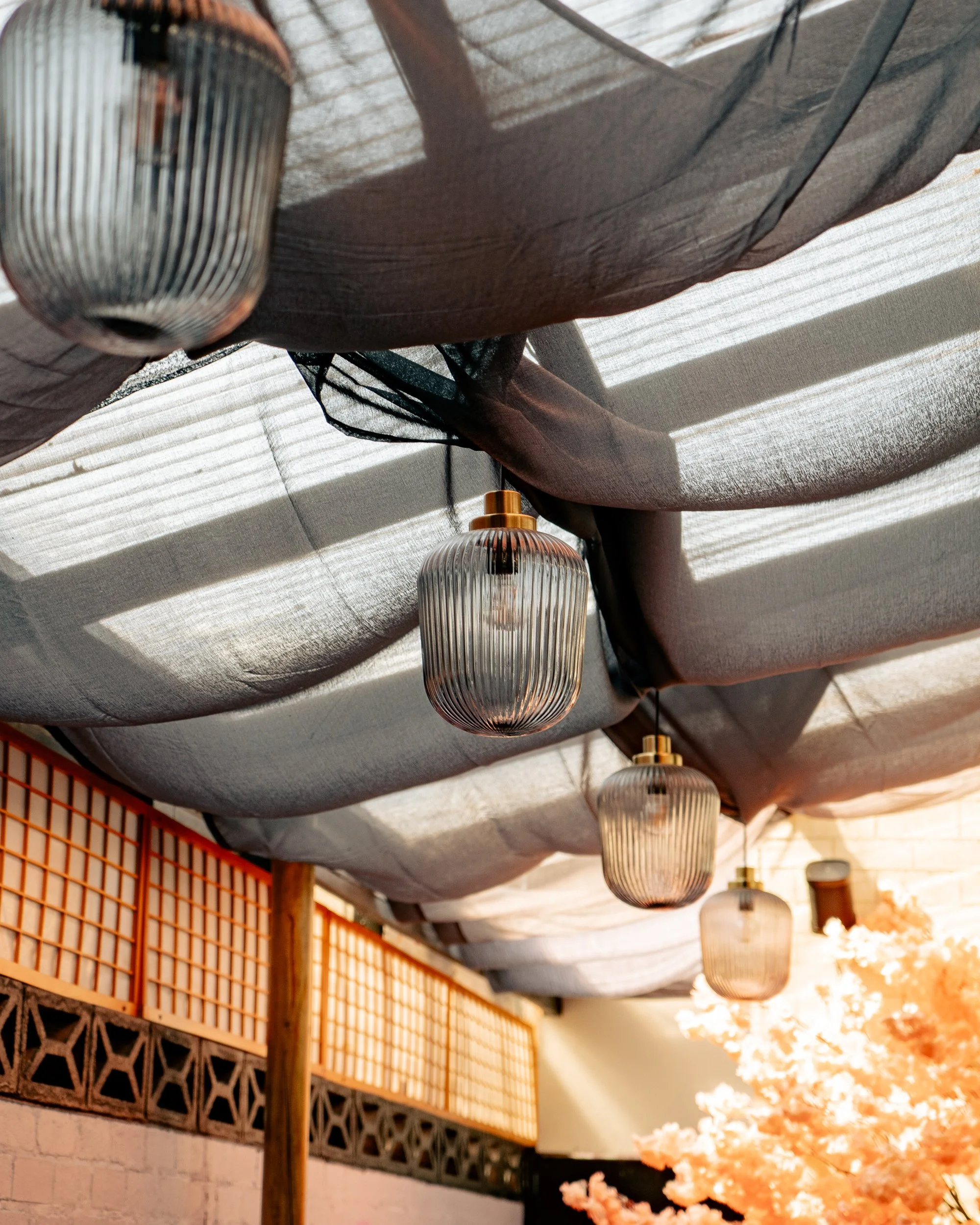 Hanging glass lanterns with black fabric draped above, and a decorative wooden lattice against a white wall, with pink flowers in the background.