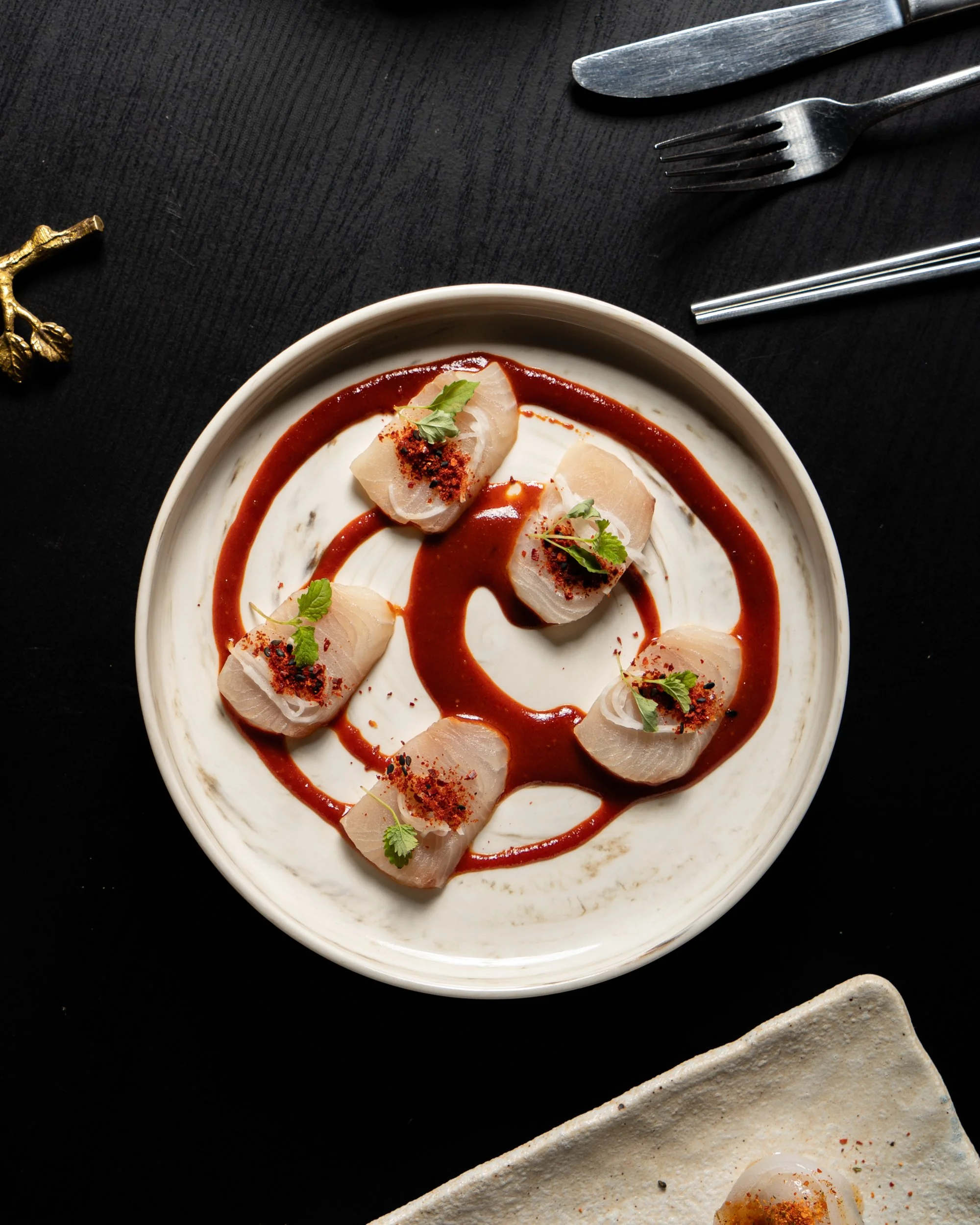 A white bowl containing five pieces of sashimi topped with herbs and spices, garnished with a dark sauce and a creamy white sauce underneath, on a black surface with cutlery in the background.