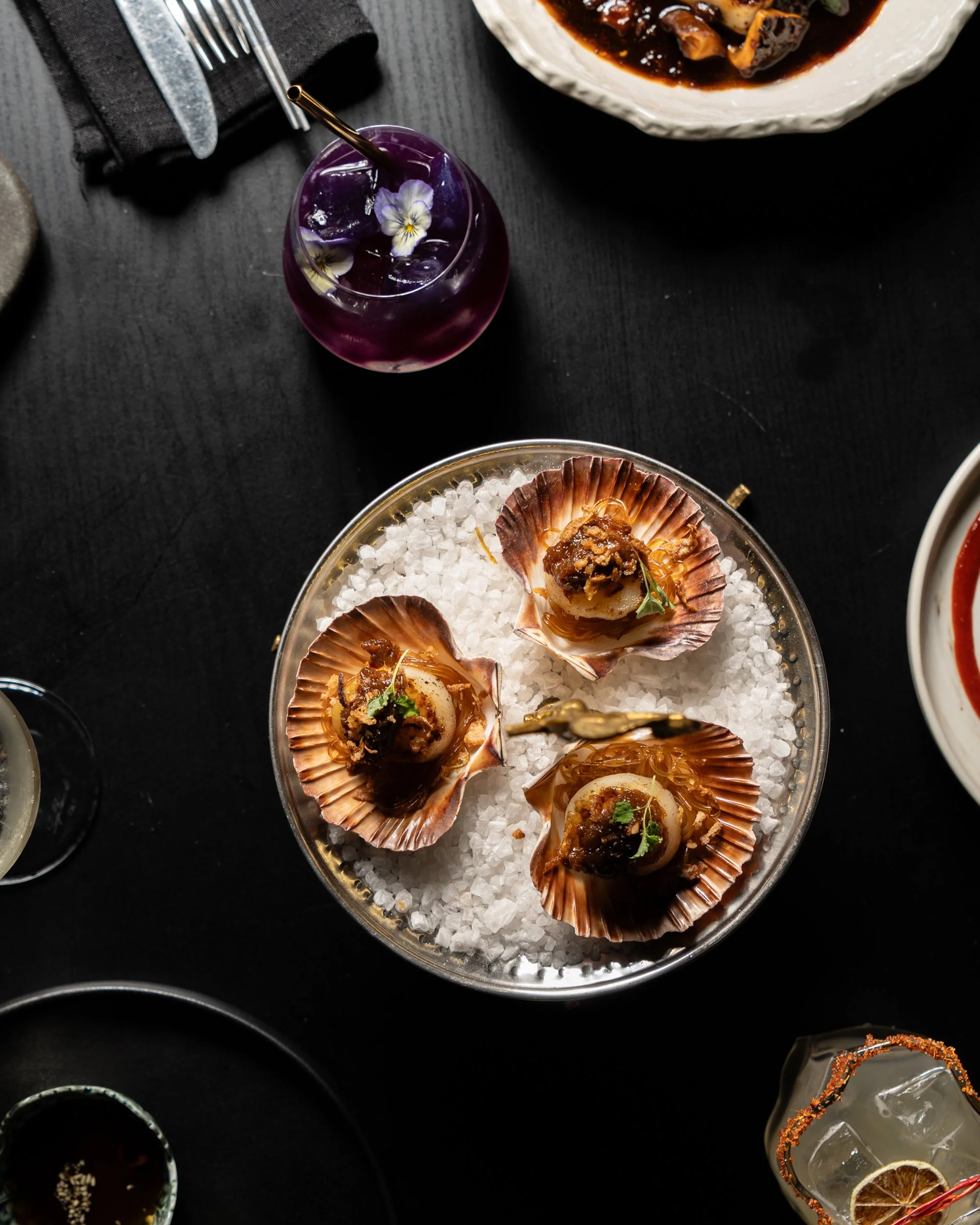 A glass bowl filled with sea urchin shells containing seafood, served on a bed of coarse salt, with a purple drink garnished with flowers nearby.