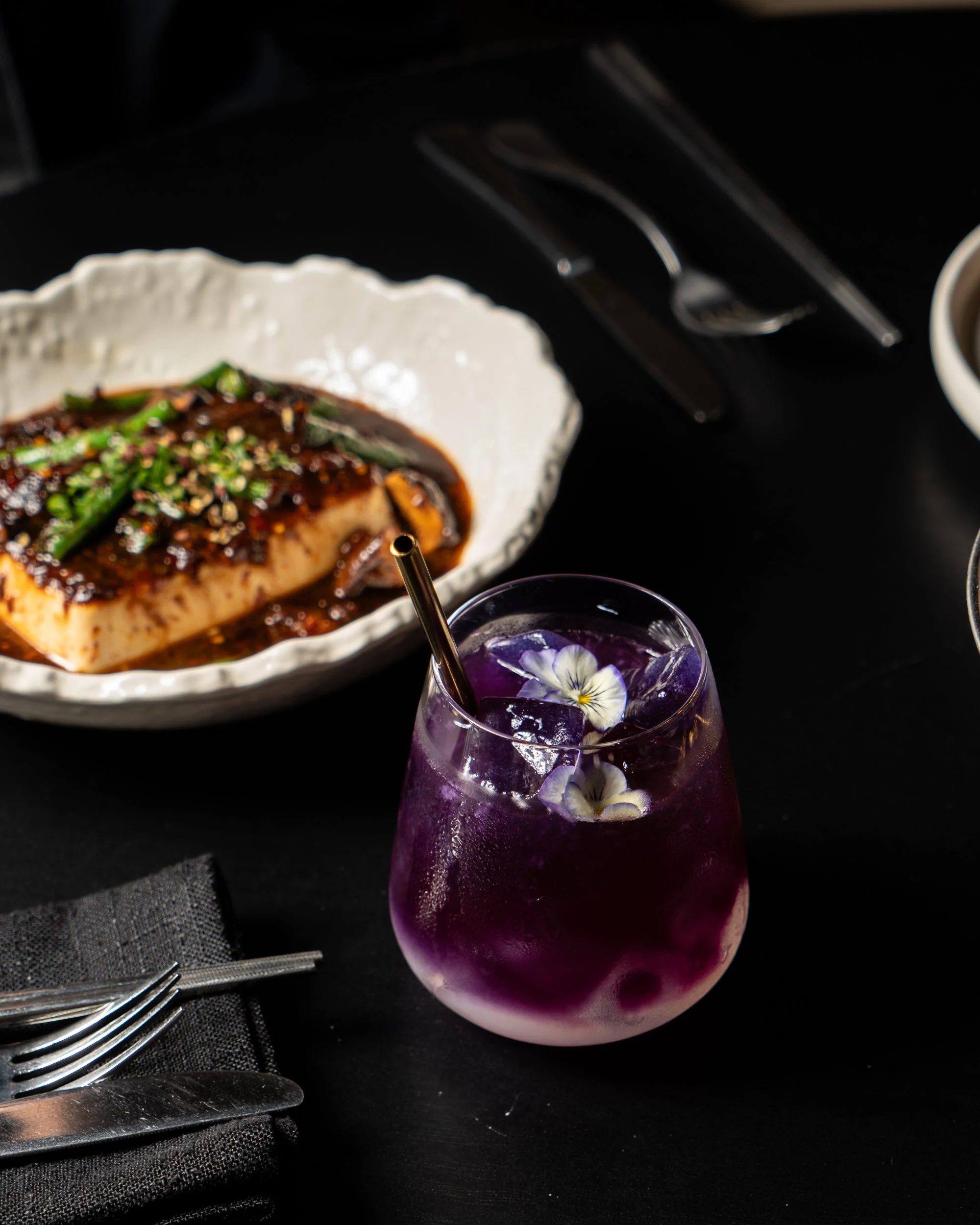 A glass of purple cocktail with ice and edible flowers, along with a dish of tofu topped with sauce and green onions on a black table.