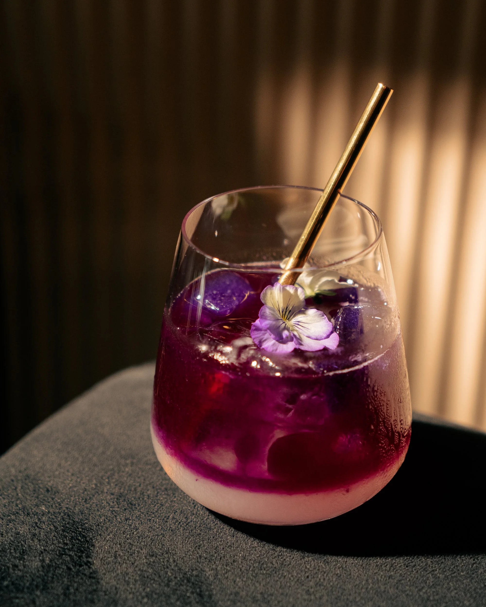 A glass of purple and pink layered cocktail with ice cubes, garnished with a purple flower and a gold straw, on a dark surface.