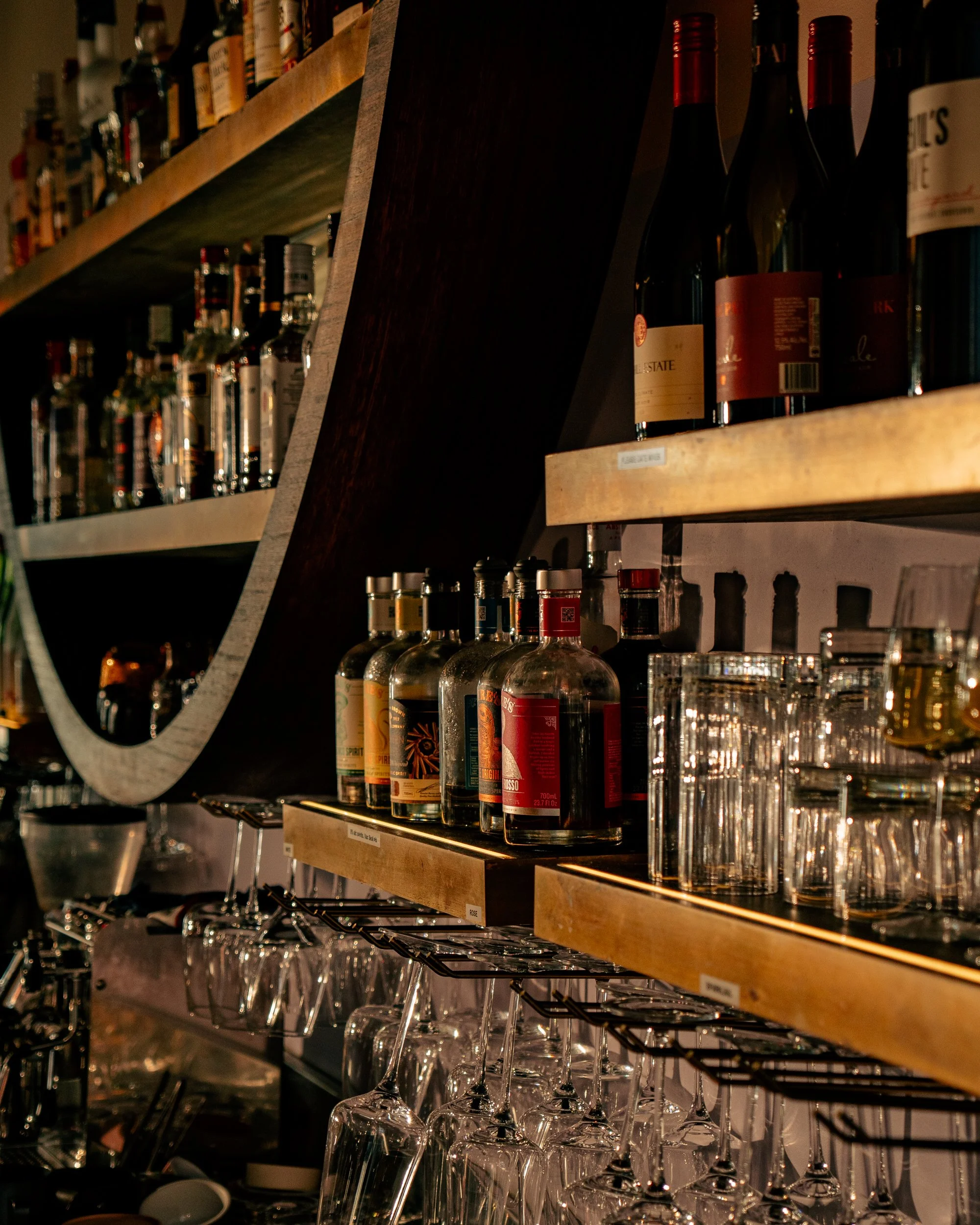Shelf with various bottles of alcohol, glasses, and barware in a dimly lit bar.