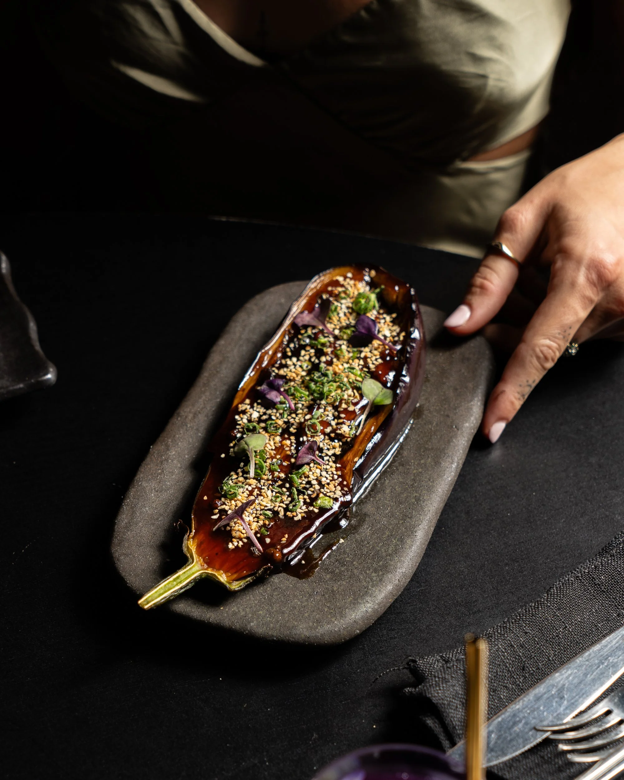 A baked eggplant dish topped with sauce, sesame seeds, green herbs, and purple microgreens, served on a dark rectangular plate.