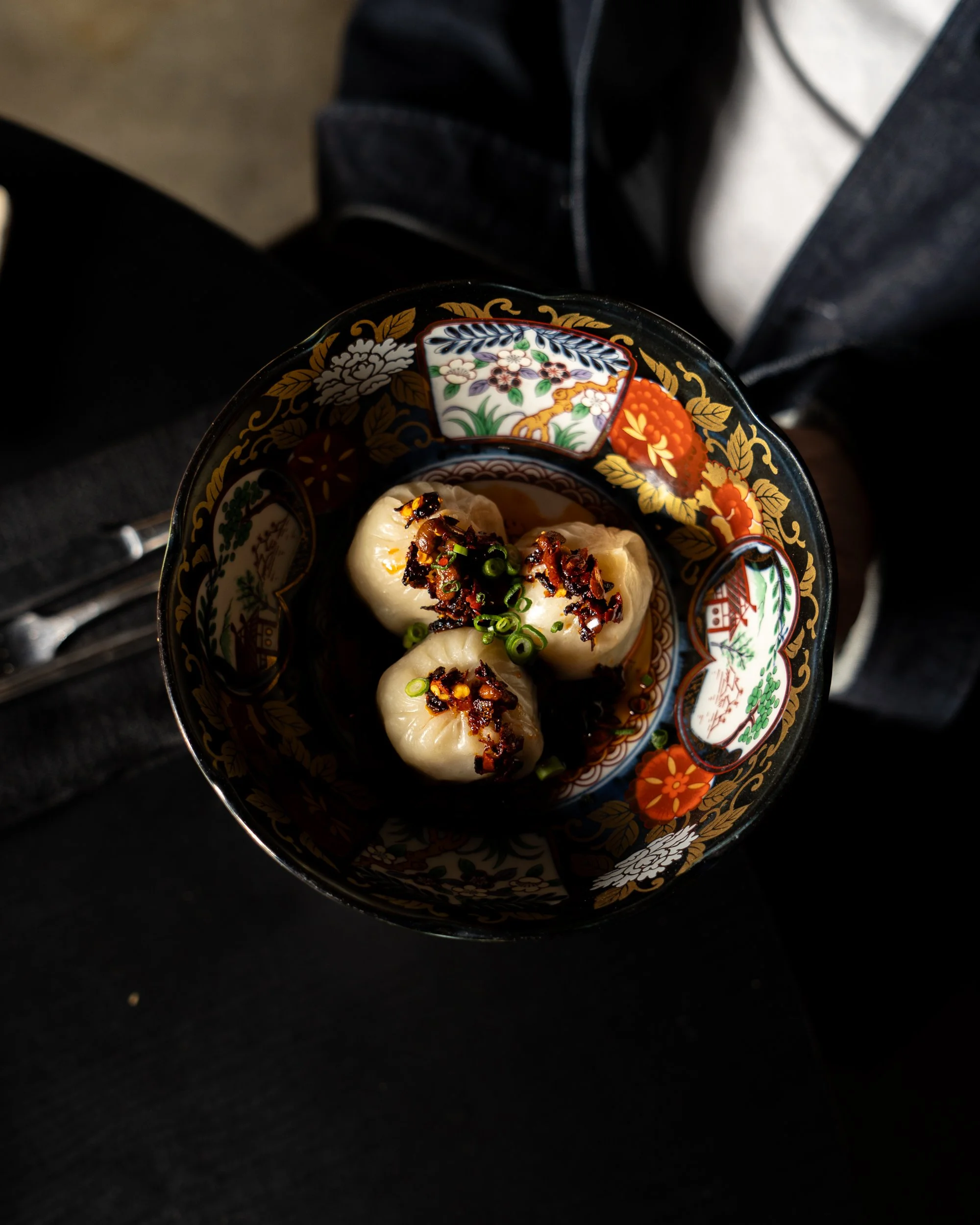 A decorative bowl containing three dumplings topped with chopped green onions and crispy fried shallots, with soy sauce in the bowl.