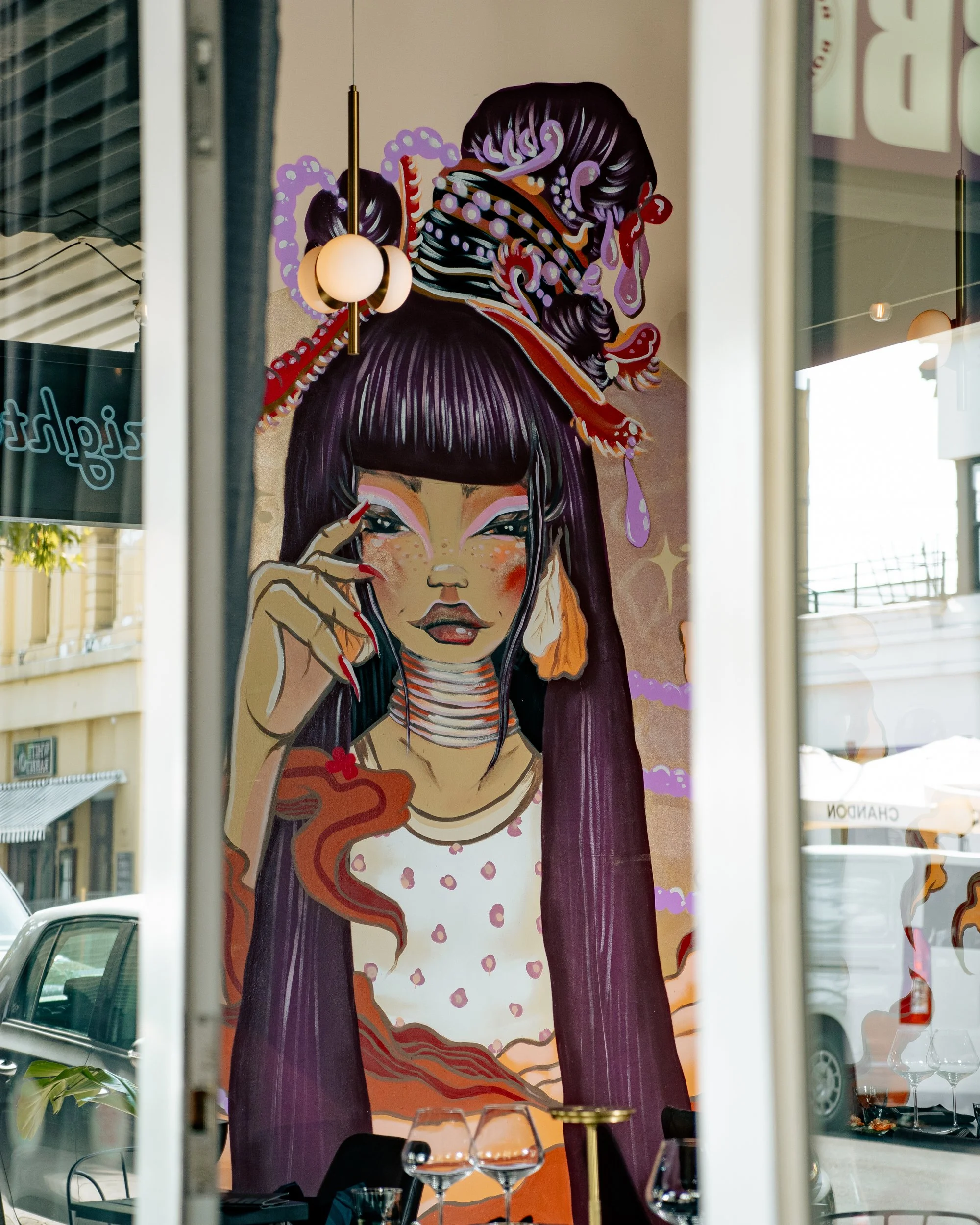 Colorful mural of a woman with long dark hair, elaborate hair accessories, bright makeup, and wearing a polka-dot dress, seen through a window reflection.