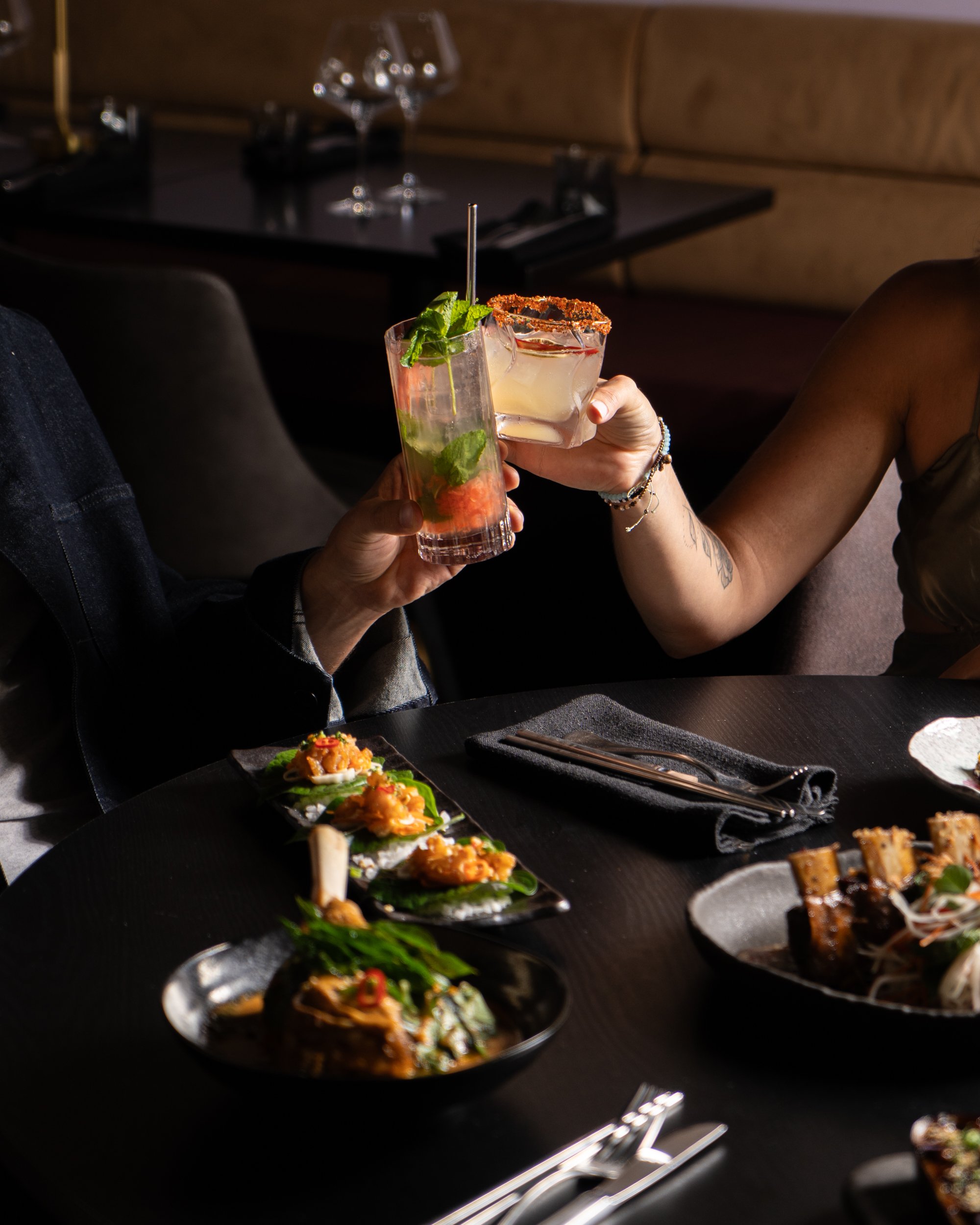Two people clinking glasses at a dinner table with various dishes, including sushi and grilled meat, in a dimly lit restaurant.