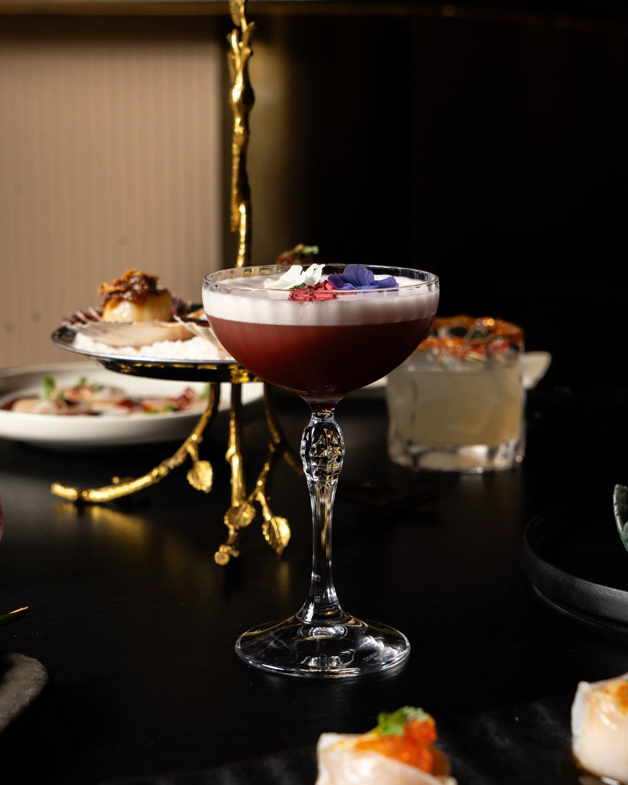 A fancy cocktail with purple, white, and red flower garnishes in a stemmed glass, on a black table with various plates of food in the background.