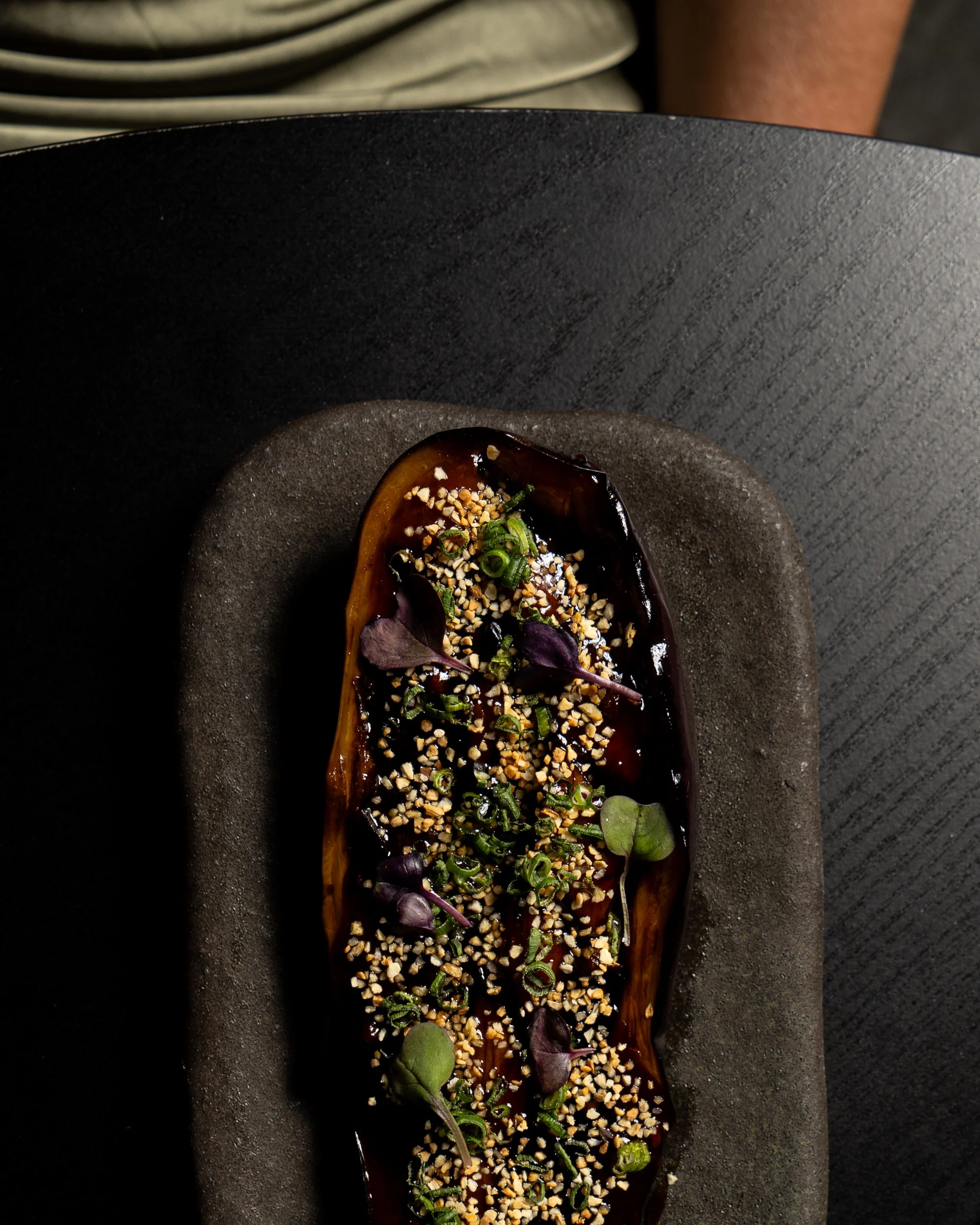 Close-up of a vegetarian dish with eggplant, topped with chopped nuts, chopped green onions, and microgreens, served on a black rectangular plate.