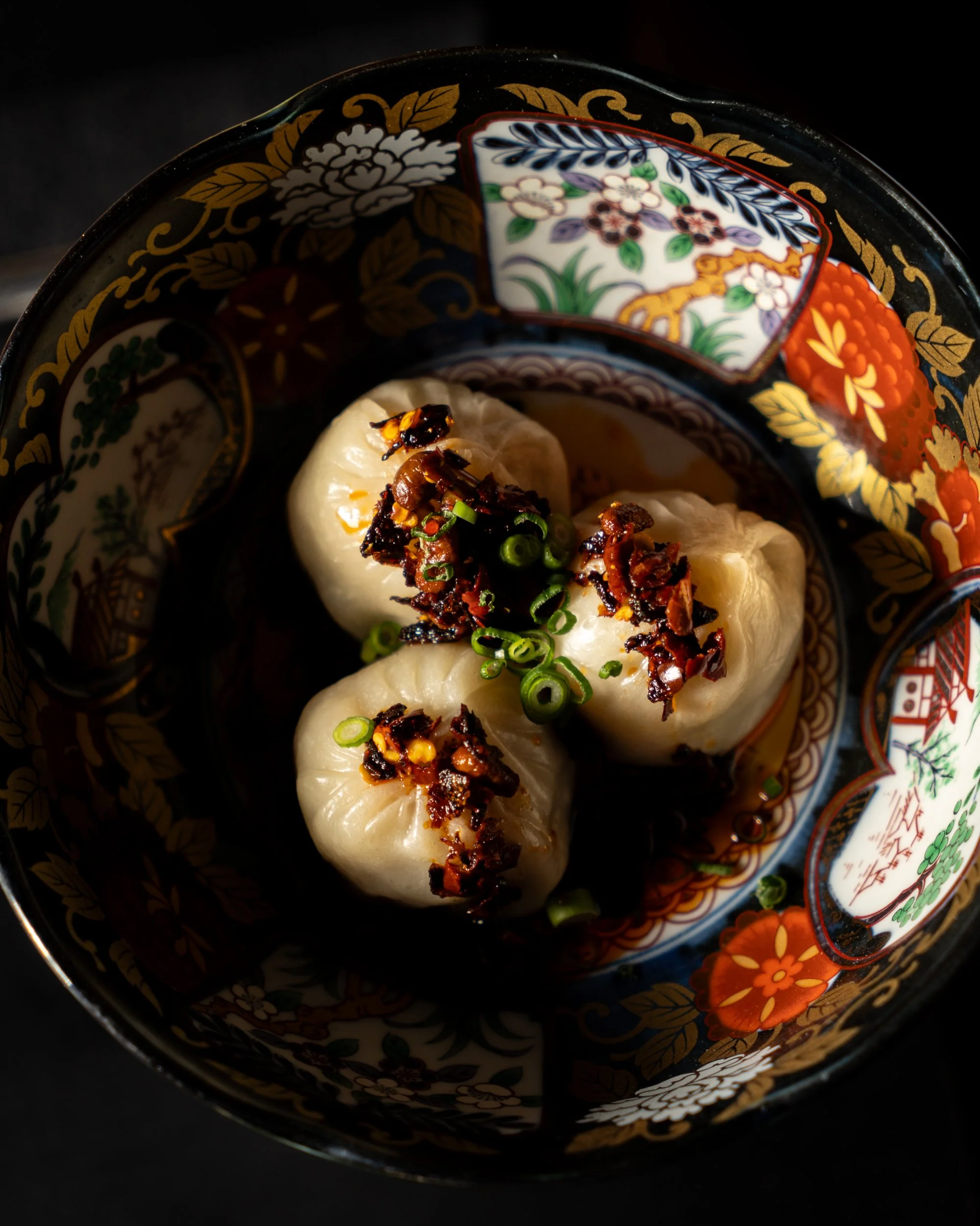 Three steamed dumplings topped with chopped green onions and dark sauce, served in a decorative bowl with floral and traditional Asian patterns.