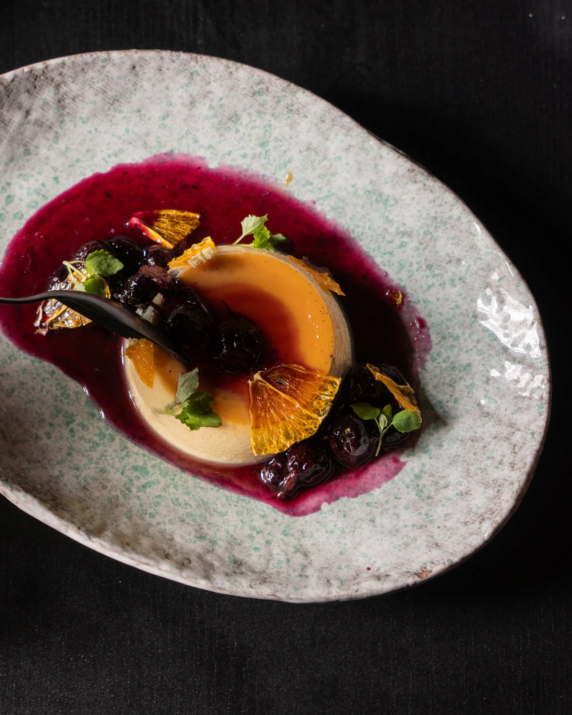 A plated dessert featuring a flan with berry sauce, fresh blueberries, orange slices, and microgreens on a decorative ceramic plate.