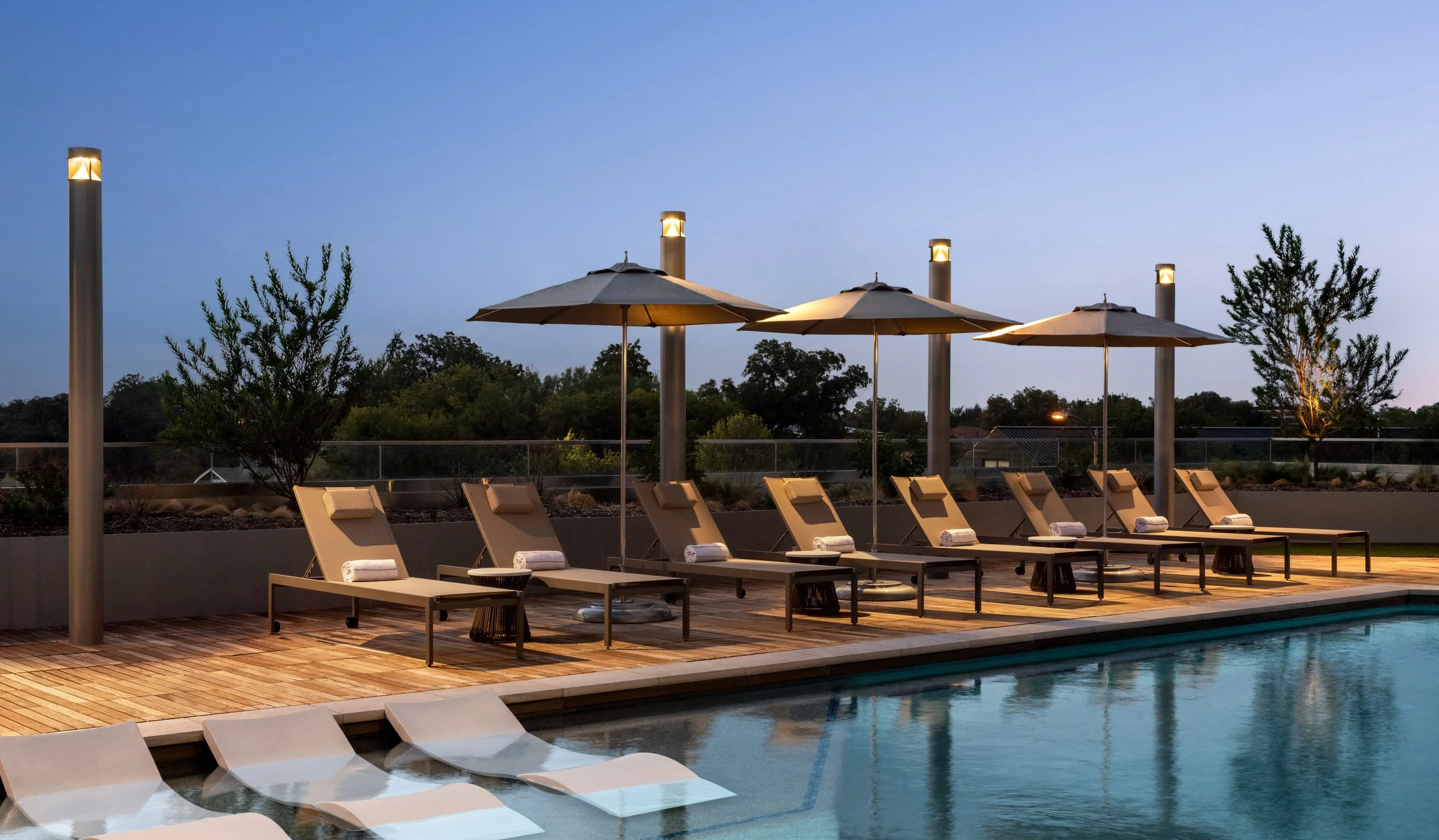 Dusk view of hotel pool with illuminated water, lined lounge beds with towels, and moody ambient lighting