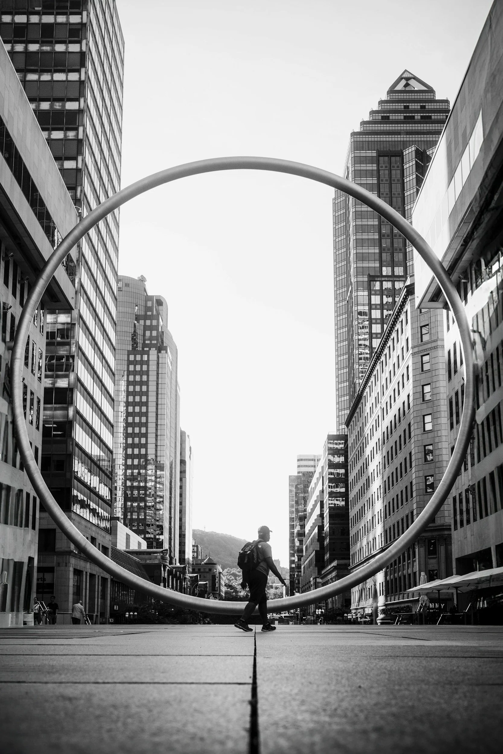 A person walking through a city street with tall skyscrapers on both sides and a large circular sculpture in the foreground, black and white photo.