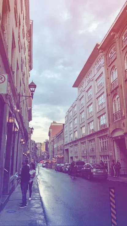 Street view in a city with buildings on each side, parked cars along the curb, and people walking on the sidewalk under a cloudy sky.