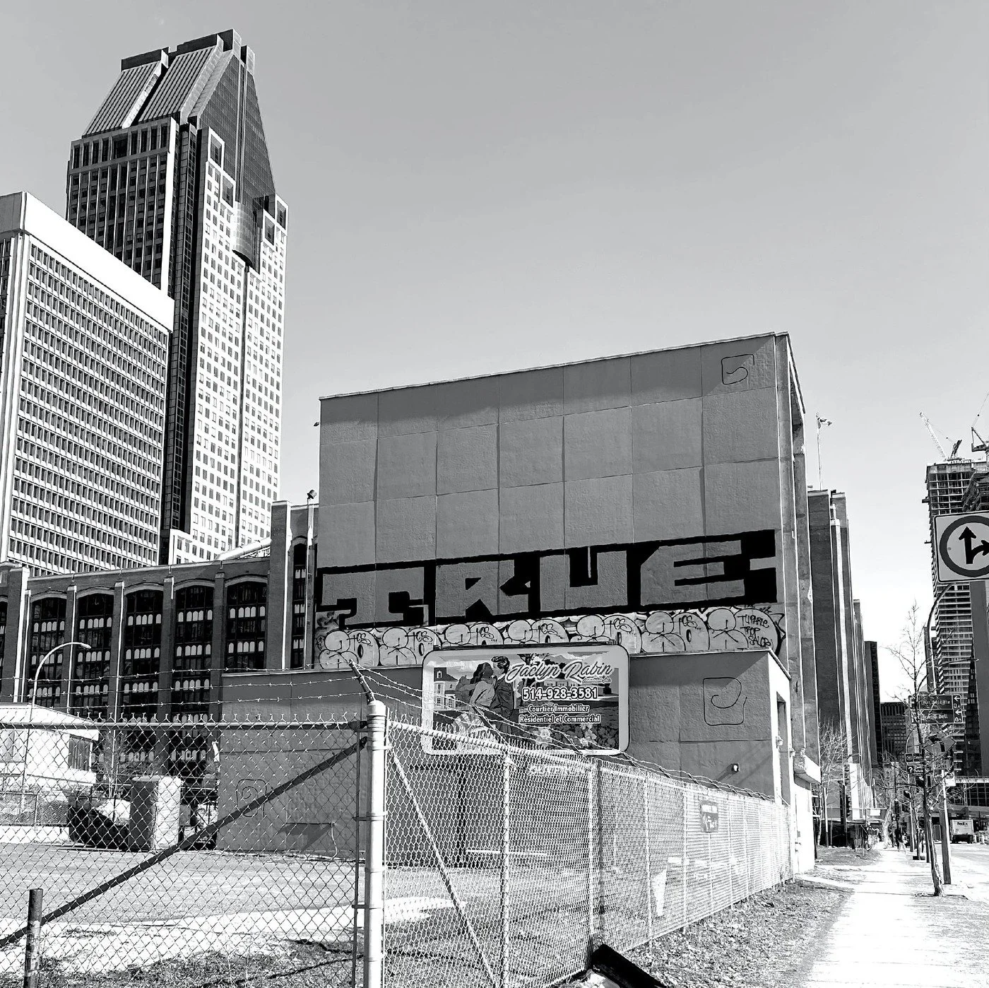 Black and white photo of a cityscape with tall buildings, including a distinctive skyscraper with a slanted top. In the foreground, a fence surrounds a construction site with a commercial building featuring graffiti art and signs, with a street visible to the side.