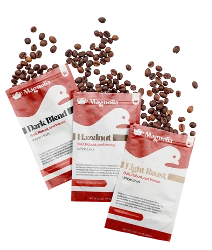 Three bags of Magnolia brand whole bean coffee labeled 'Dark Blend', 'Hazelnut', and 'Light Roast' with coffee beans spilled around them.