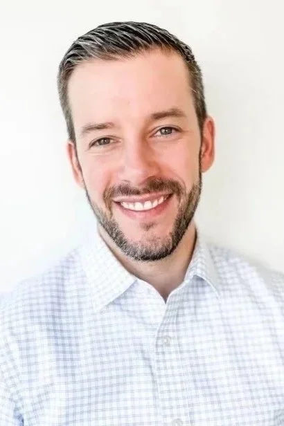 A smiling man with short brown hair and a beard, wearing a white checkered dress shirt, posing against a light background.