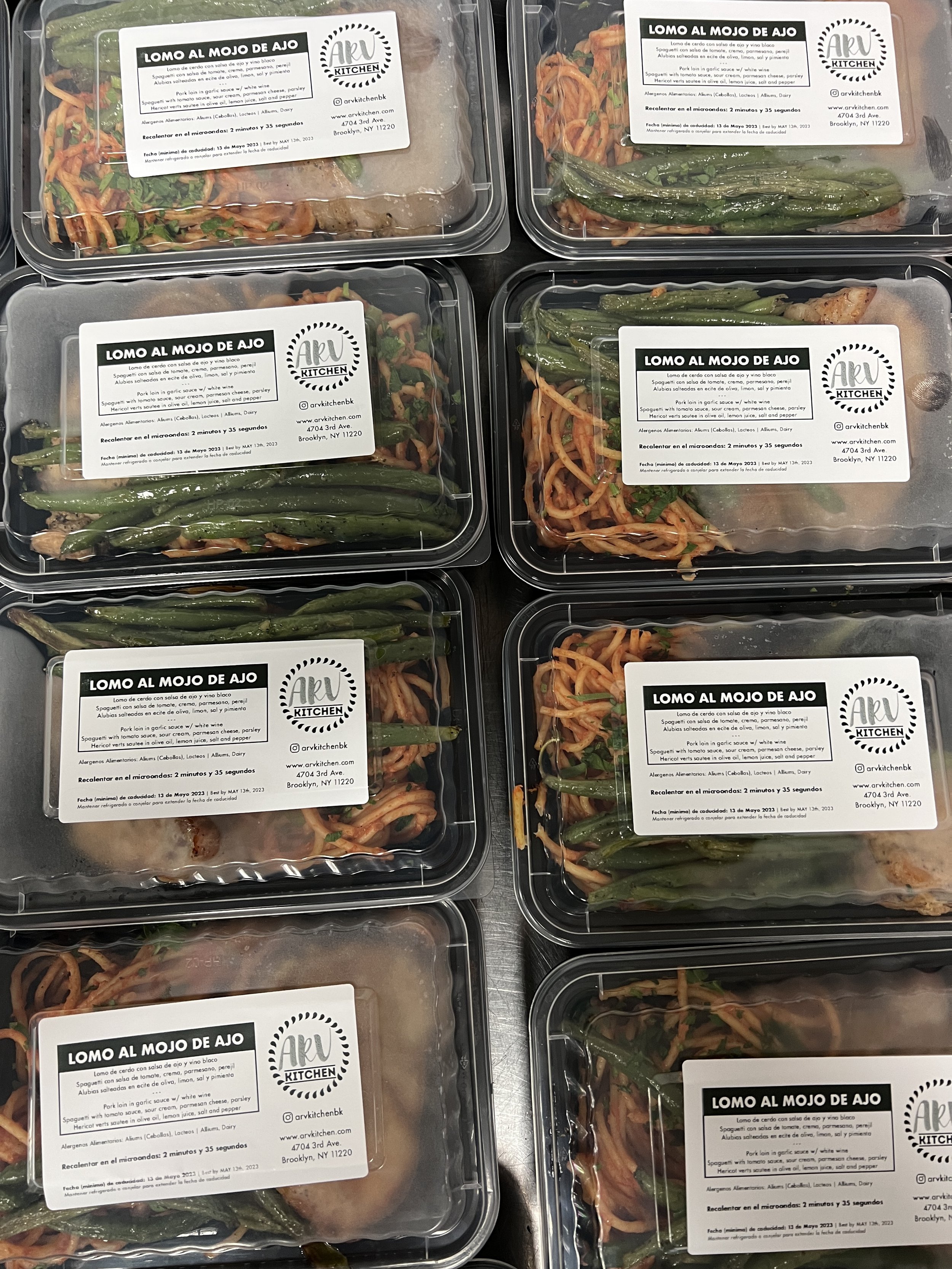 Multiple plastic containers of ready-to-eat pork loin with a side of green beans and noodles, labeled "Lomo al Mojo de Ajo" from Ark Kitchen in Brooklyn, NY.