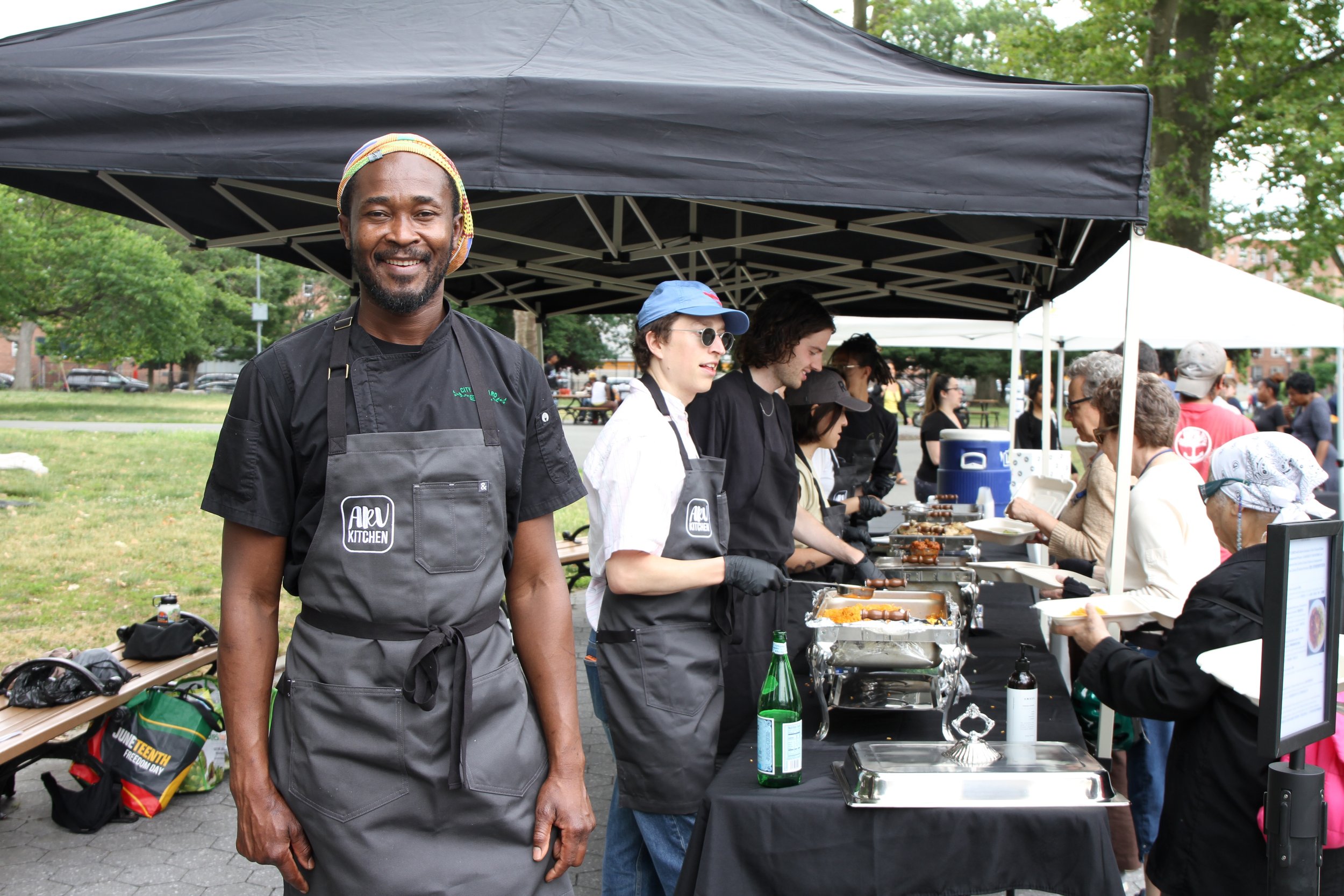 A smiling man wearing a black chef jacket and a gray apron, standing at a food stall during an outdoor event, with several people serving food behind him under tents.