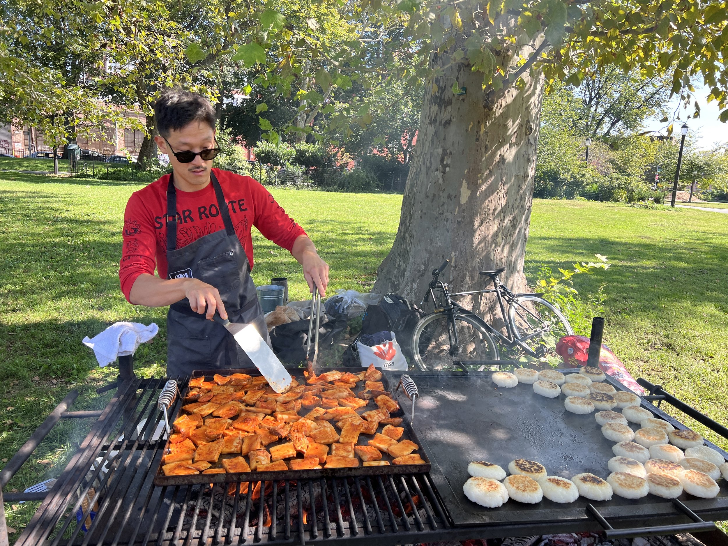 A man grilling sliced meat and bread rolls on outdoor grills in a park, with trees, grass, bicycles, and a fence in the background.