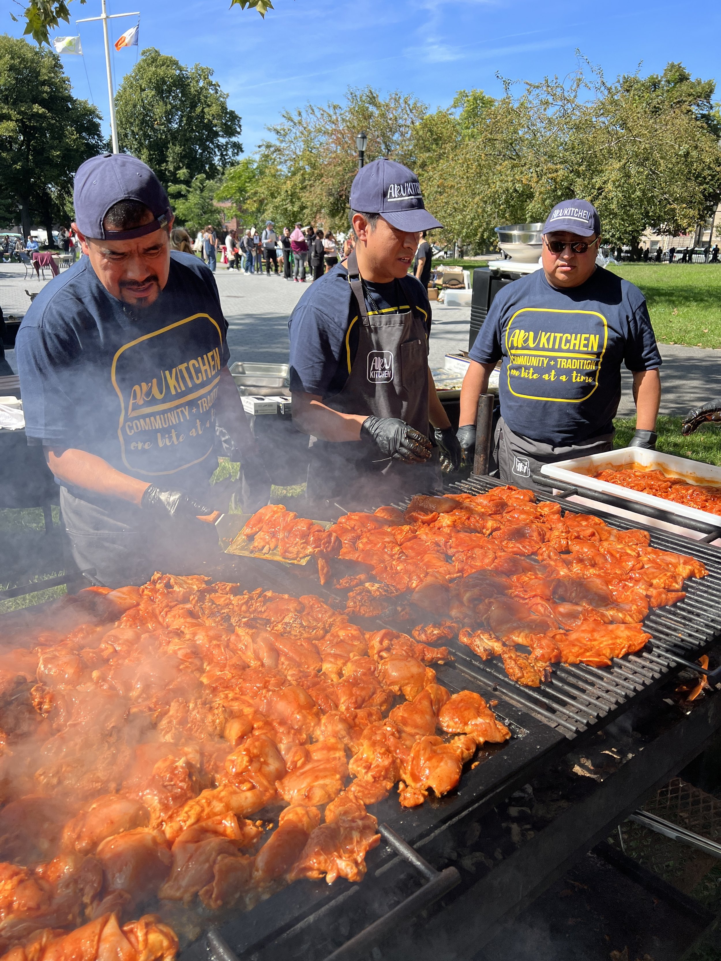 Three men cooking marinated chicken on a large outdoor grill at a community event
