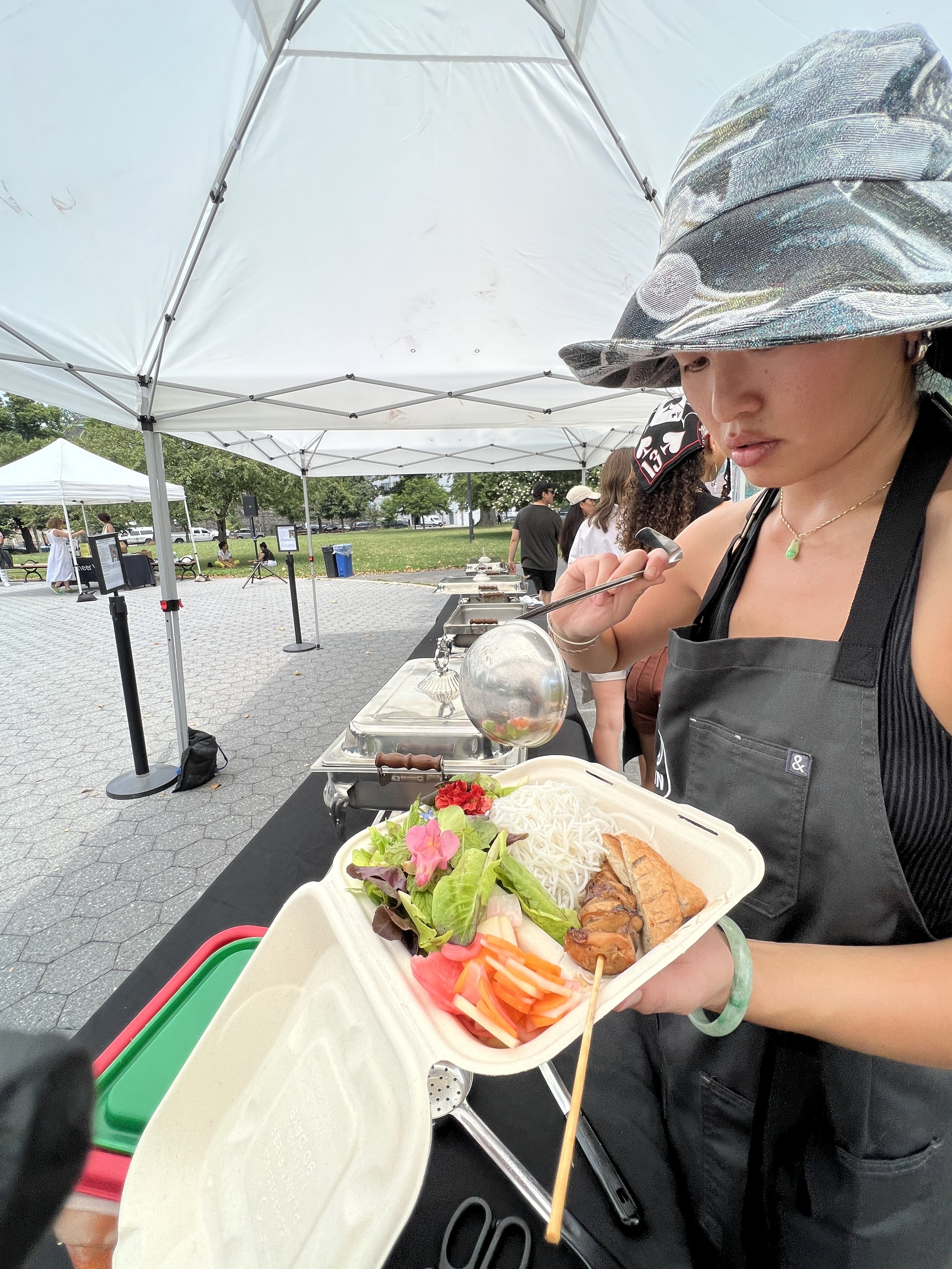 A woman wearing a camouflage bucket hat and a black apron holding a takeout container with noodles, grilled chicken, salad, and vegetables at an outdoor food event under a white canopy.