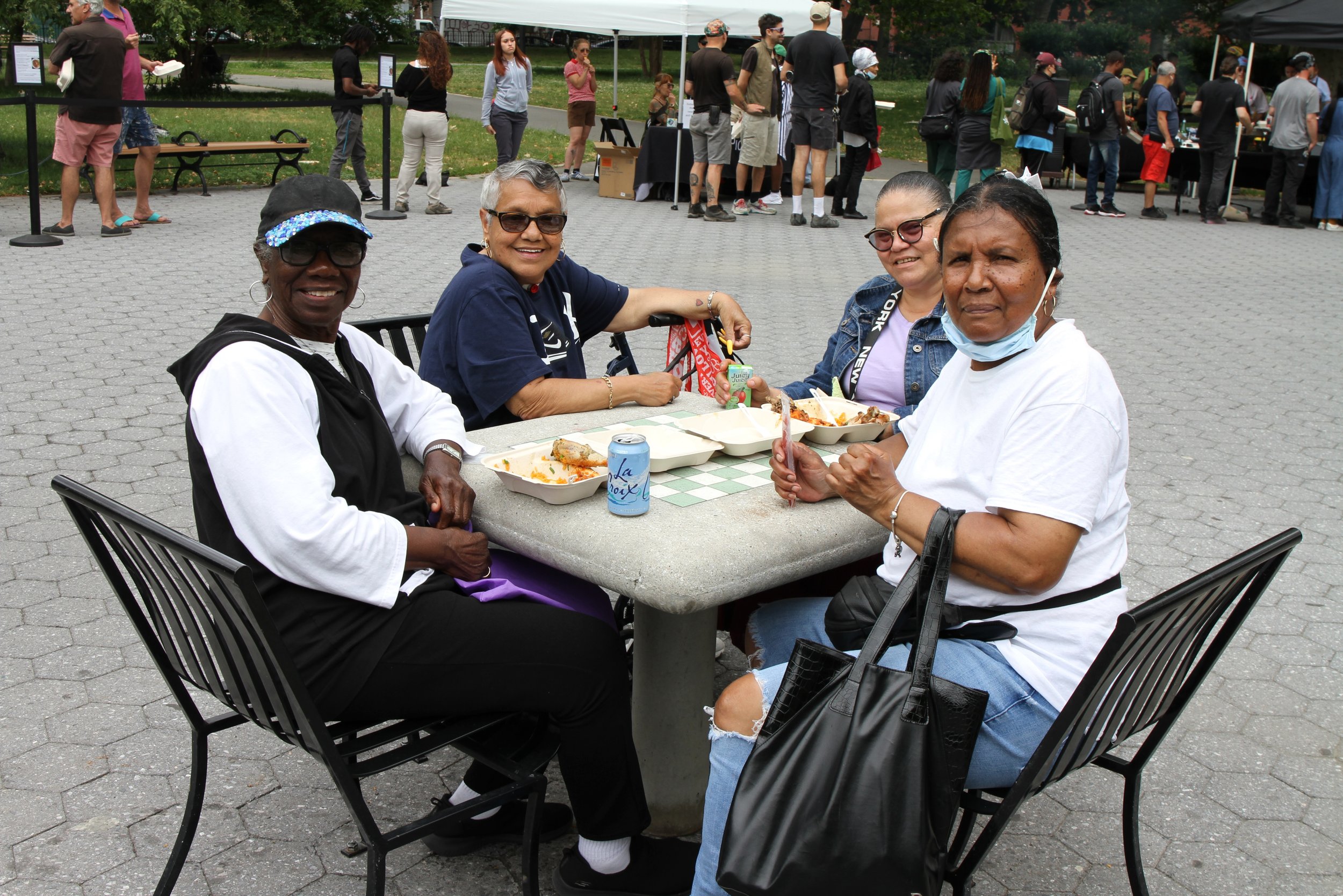 Four women of diverse ethnicities sitting at an outdoor table eating food at a community event, with a crowd of people standing in line in the background.