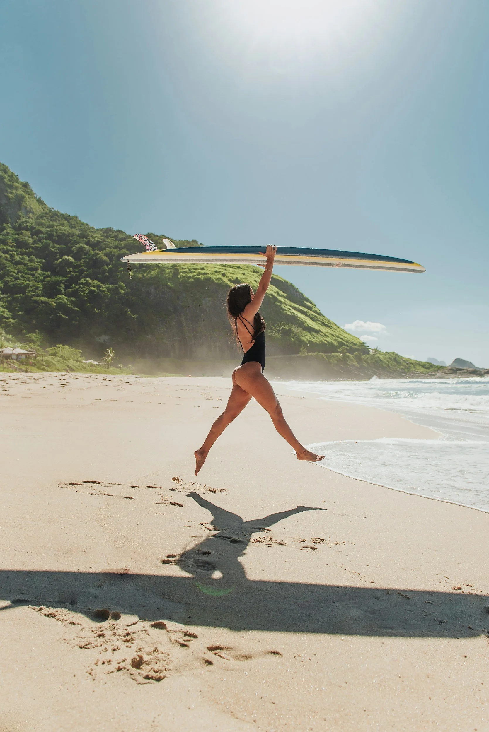 A woman in a black swimsuit jumping on the beach with a surfboard overhead, with a green mountain and clear blue sky in the background.