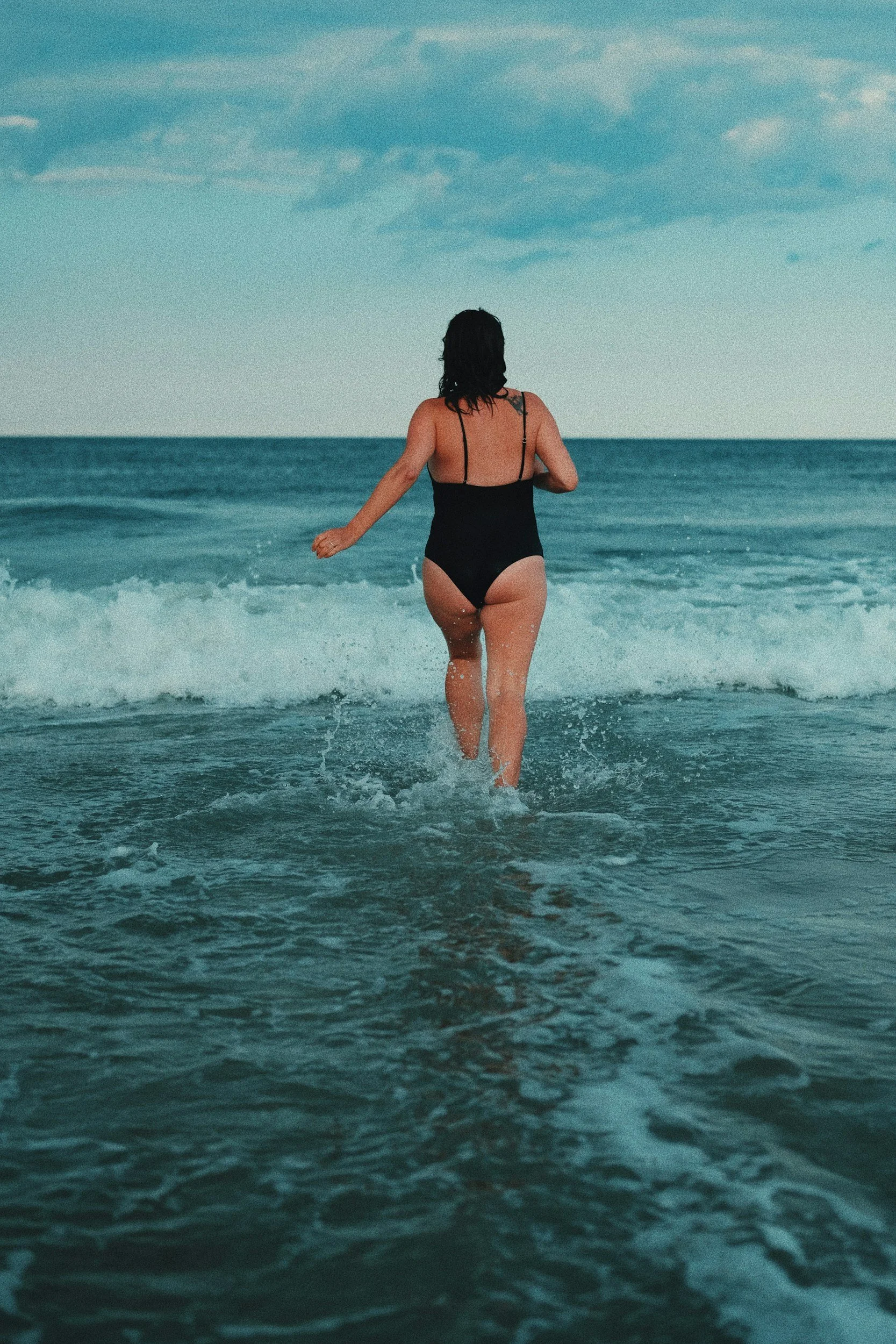 A woman in a black swimsuit walking into the ocean with her back facing the camera, waves around her legs, under a partly cloudy sky.