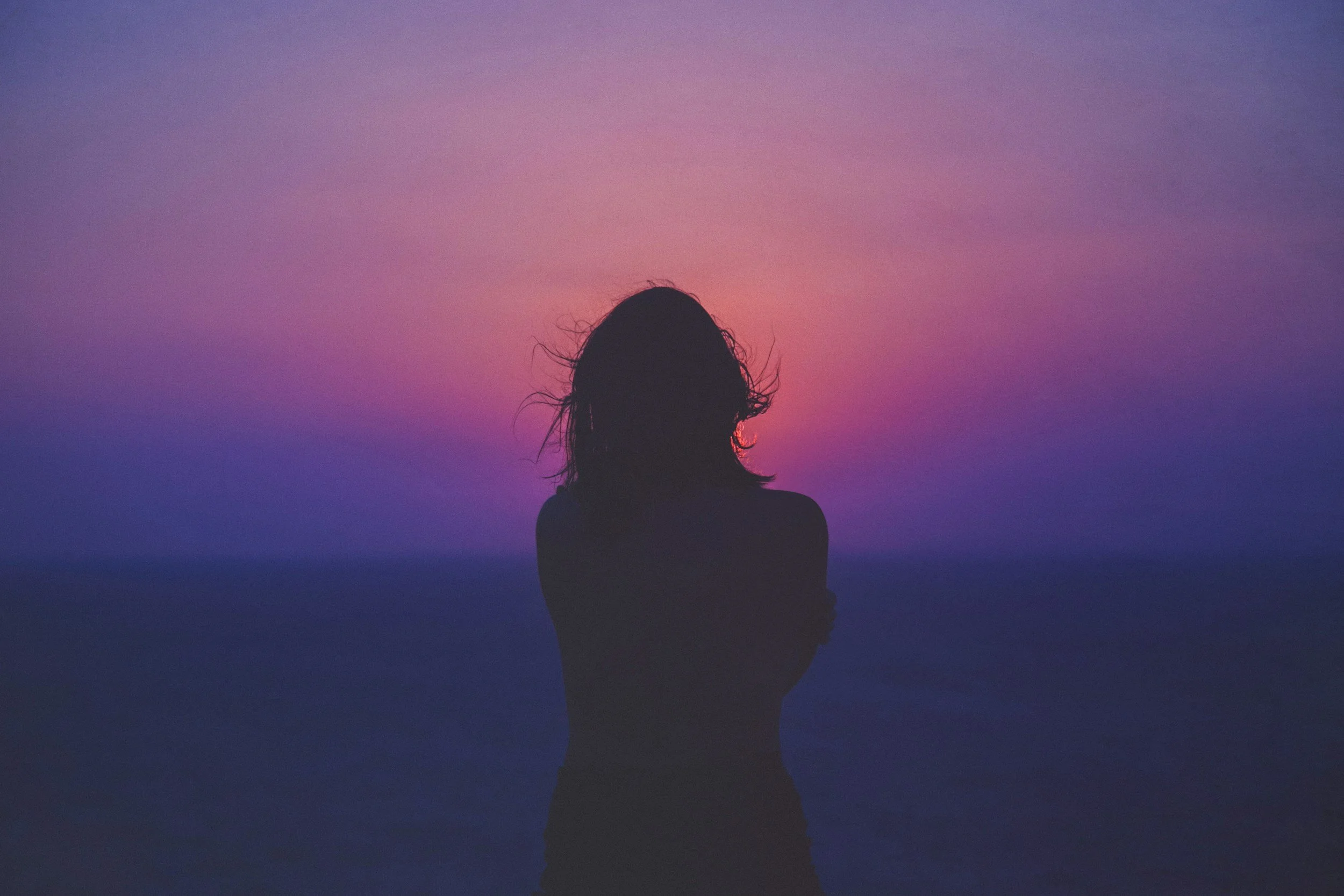 Silhouette of a person with messy hair standing with crossed arms at sunset or sunrise over a body of water, with colorful purple and pink sky.