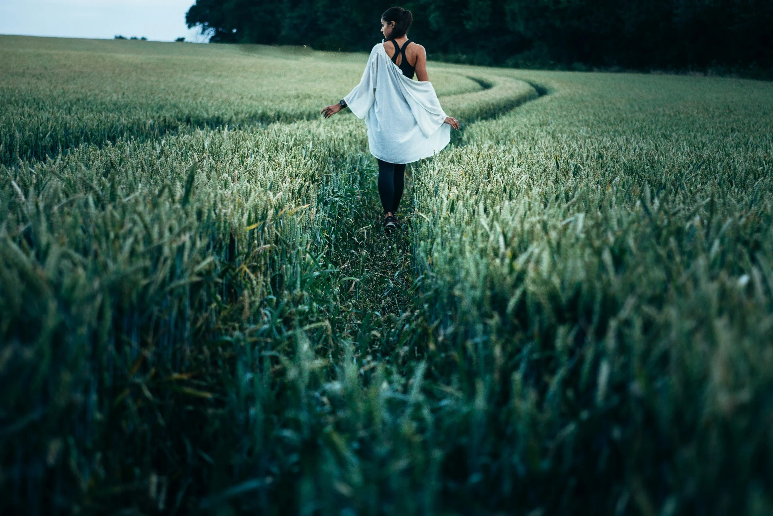 A woman in a black outfit with a white shawl walking through a green wheat field.