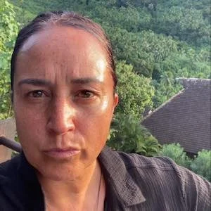 Close-up of a woman with dark hair pulled back, outdoors with green trees and a house roof in the background.