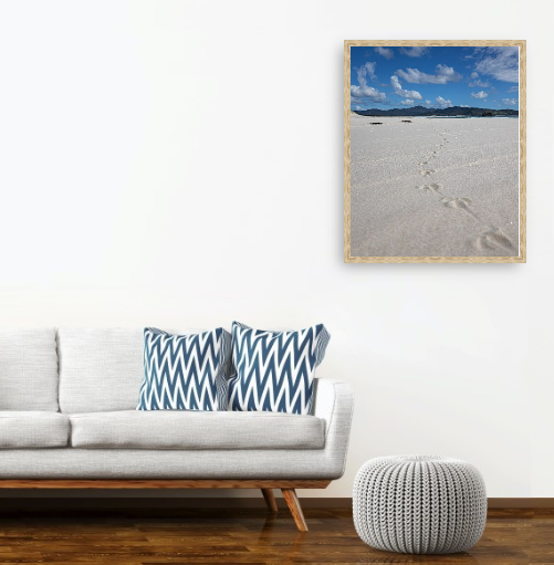 Modern living room with white sofa, chevron-patterned pillows, round ottoman, and wall art of beach scene with footsteps.